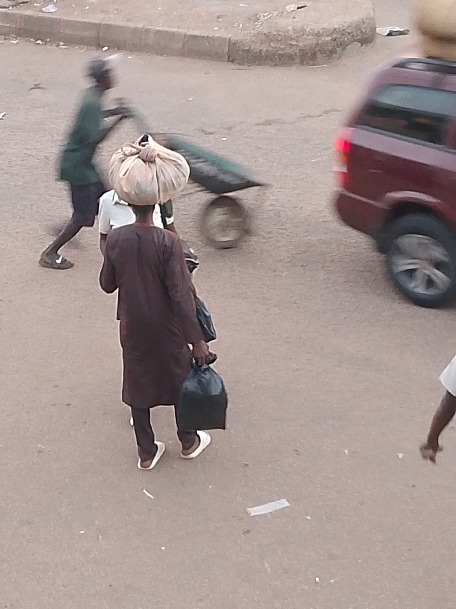 In Bukuru, Nigeria, a scene unfolds on a paved road. A person is walking away from the camera, carrying a large, light-colored bundle balanced on their head, covered by a bag tied at the top. They are wearing a dark, long-sleeved garment and dark trousers. In their right hand, they are holding a black plastic bag. To their left, another person, partially visible, is wearing a white shirt. Further to the left, a person in a green long-sleeved shirt and dark shorts is pushing a cart. The cart appears to be carrying cylindrical objects covered in a green material. A maroon minivan is visible on the right side of the frame, moving. The ground is a dusty, light brown pavement. The overall lighting suggests it is daytime.
