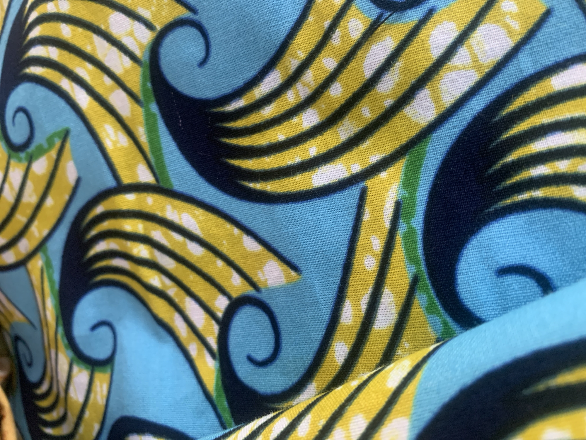 The image is a close-up, overhead shot of a vibrant blue fabric with a bold, repeating pattern. The pattern consists of stylized, curving shapes in bright yellow and dark navy blue, outlined by thin black lines. Scattered within the yellow shapes are small white dots. There are also thin green accents interspersed within the design. The fabric appears to be made of a woven material, with visible threads creating a textured surface. The way the fabric is folded and creased suggests it might be clothing, a textile for sale, or a decorative item. No specific location in Jauro Yinu, Nigeria can be determined from the image itself as it focuses solely on the fabric's pattern.