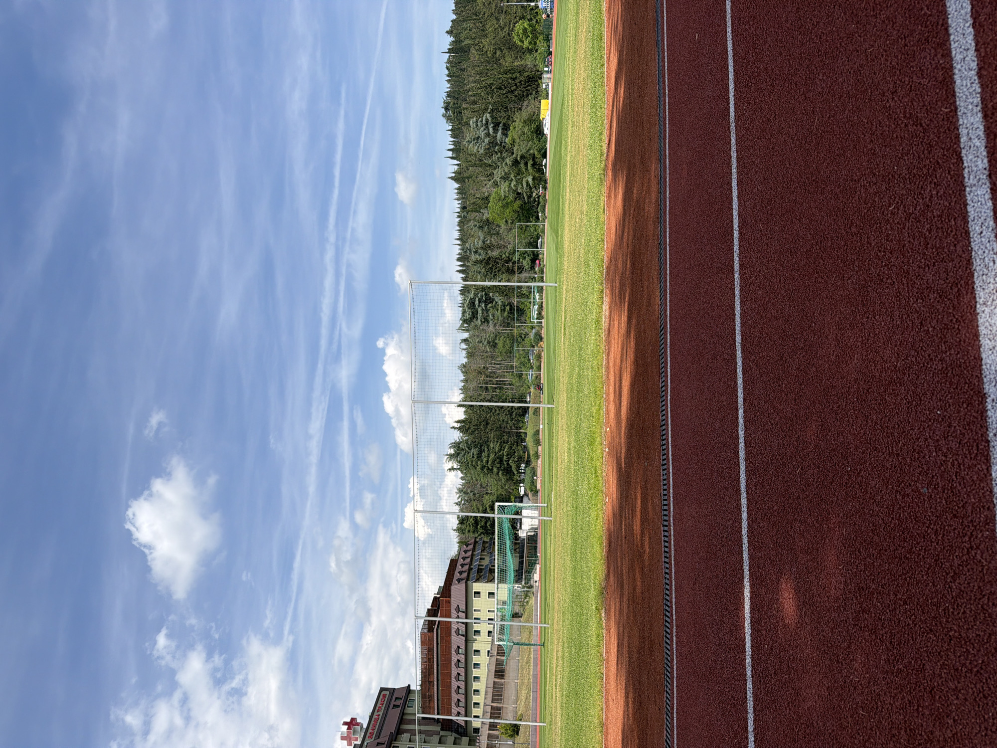 An outdoor sports complex is depicted under a bright blue sky with scattered white clouds and visible contrails. The image captures a red athletic running track with crisp white lane markings in the foreground and along the right side. Shadows are cast onto the track, suggesting sunlight from the left.
Adjacent to the track is a vibrant green grass field, resembling a football or soccer pitch. A white goalpost with a green net is visible on the field in the midground, behind which a wire mesh fence extends across the scene.
In the background, a dense line of tall, dark green trees forms a natural screen. To the left, behind the trees and fence, a large multi-story building with sloped roofs, balconies, and numerous windows is visible. On this building, a prominent red cross symbol and some red, partially legible text are present. Given the location context of Stara Zagora, Bulgaria, and the medical cross symbol, this text likely refers to a hospital or medical facility, possibly "Traiana Hospital."
The scene is devoid of people, presenting an empty sports facility ready for use. The clear sky and bright illumination indicate good weather during daytime, likely late morning or early afternoon.