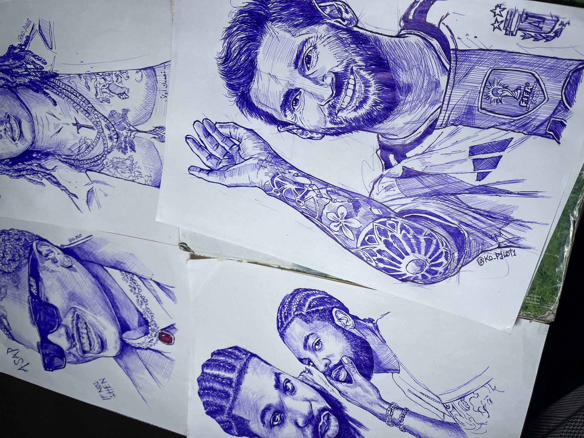 The image is a close-up, overhead shot of four pen and ink drawings on white paper, arranged somewhat overlapping. The drawings depict stylized portraits of several individuals.
The top left drawing shows a close-up of a person wearing sunglasses, a suit jacket, and a prominent necklace with a large red pendant. The name "ASAP" is written above the person's head, and "IF NOT WHEN" is written to the left.
The top right drawing is a partial portrait focusing on the chest and neck area of a person with dreadlocks, adorned with multiple necklaces and tattoos on their chest. Arabic script is visible below the chest.
The bottom left drawing features two individuals. The upper figure has braided hair and is looking towards the viewer. The lower figure, with similar braided hair, is shown with a hand near their mouth, appearing to speak or whisper. They are both wearing necklaces, and the lower figure has a beaded bracelet.
The bottom right drawing is a portrait of a man with short hair and a beard, identified as Lionel Messi. He is wearing a soccer jersey with the FIFA World Cup 2022 badge. His left arm is raised, and it displays intricate tattoos. The artist's signature, "@KO_PILOTI", is visible on this drawing and also faintly on the top left drawing.
The drawings are placed on what appears to be a desk or table. A corner of a green and white publication is visible at the bottom, with text such as "THE REVOLUTION DUBAI V MARINA M". The context suggests this is an artist's workspace. The request to locate this scene in Jos, Nigeria, is not supported by any visual cues within the image.
