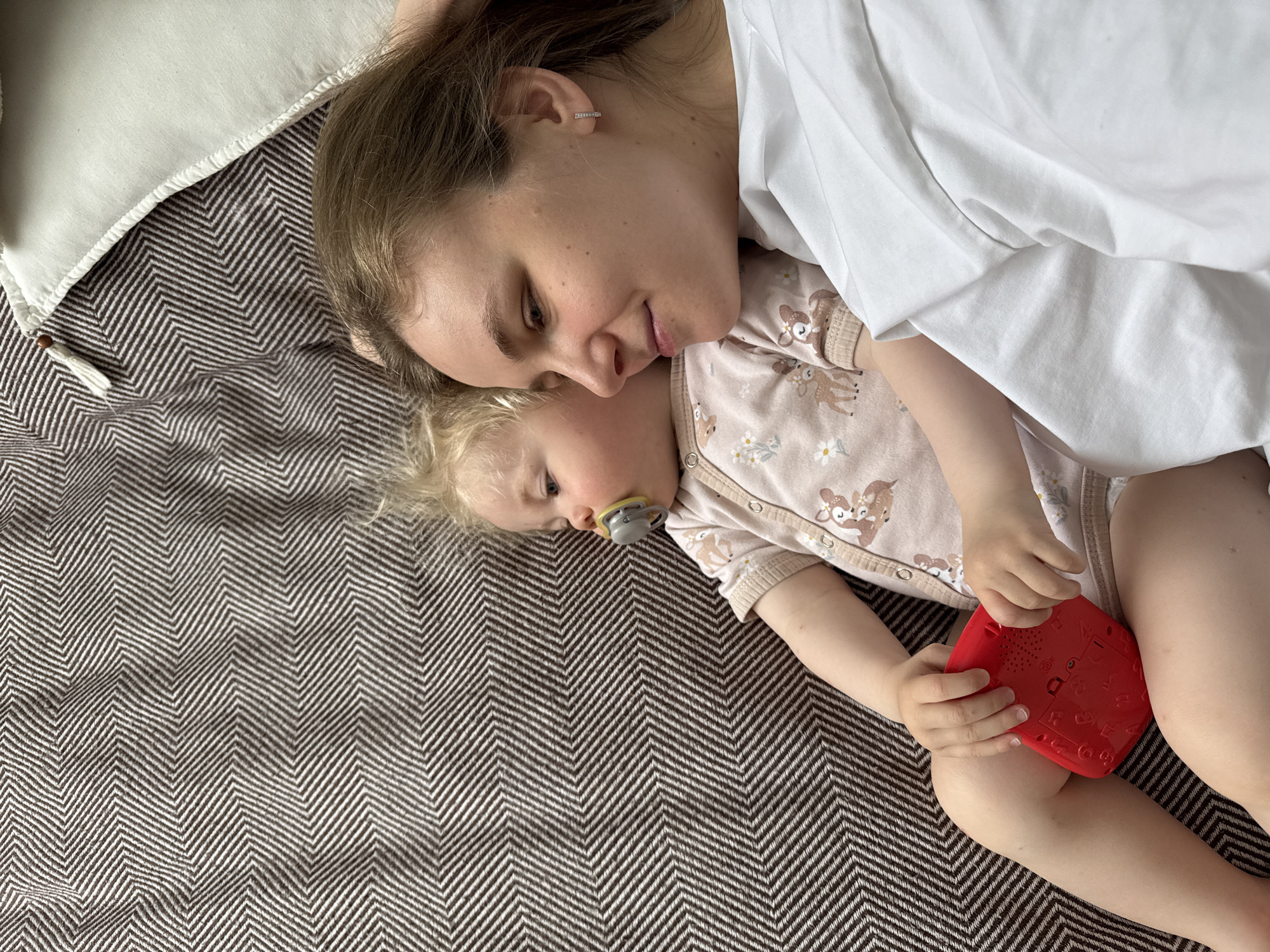 A top-down shot shows a woman and a young child lying on a brown and white chevron-patterned blanket. The woman is lying on her side with her head near the top left corner of the frame. She has light brown hair pulled back and is wearing a small earring. She looks down at the child with a soft expression. The child is lying on the blanket next to the woman, facing upwards and slightly to the left. The child has blonde hair and a pacifier in their mouth. The child is wearing a light pink onesie with an animal print. The child is holding a red cube-shaped toy in their hands. The setting appears to be indoors. The location is not discernable from the image and cannot be determined to be Vladimir, Russia.