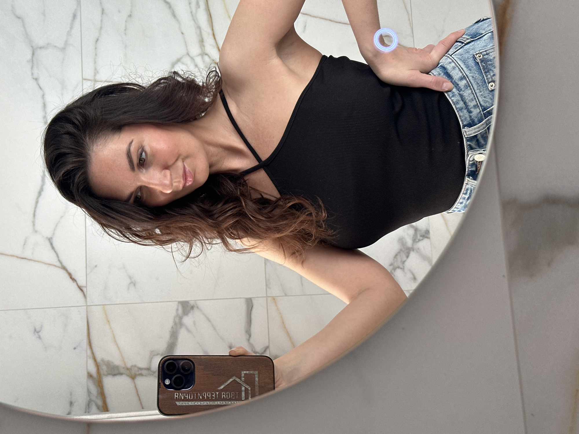 A woman is reflected in a round mirror, holding a smartphone. She is wearing a black top and blue jeans. The mirror is mounted on a wall tiled with marble-patterned tiles. The smartphone has a wooden case.
There is no indication that the location is Chaikovsky, Russia.