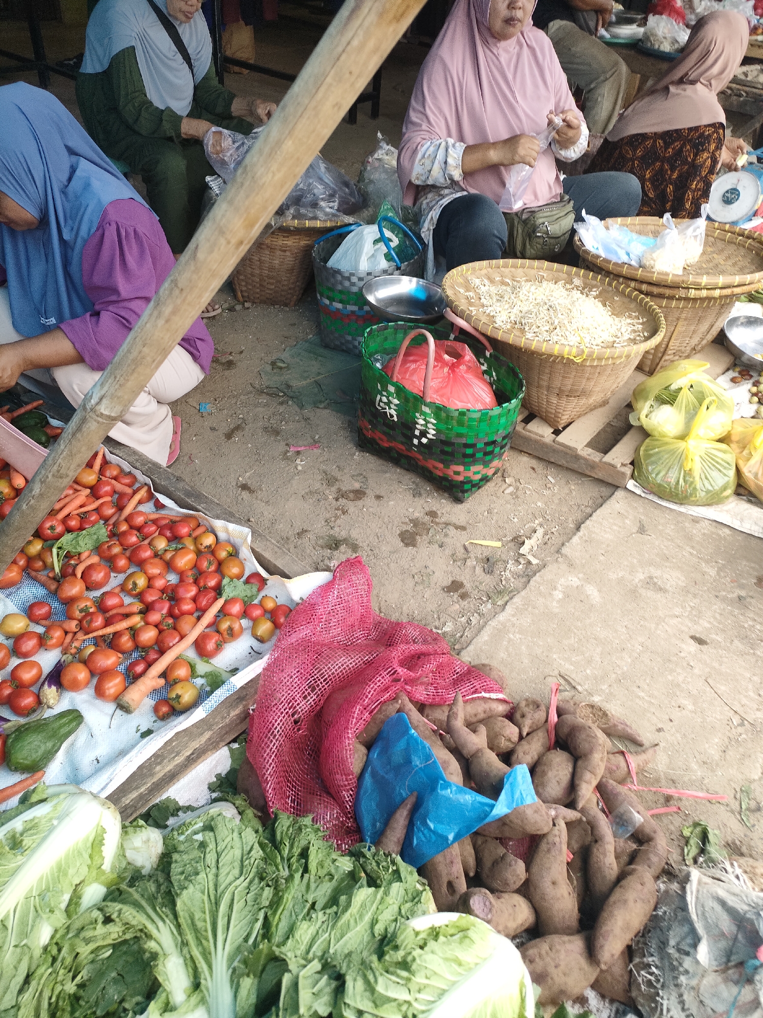 The image captures a market scene in Sukoyoso, Indonesia. Two women are visible, both wearing headscarves. The woman on the left is wearing a purple headscarf and a purple top, leaning forward and appearing to be sorting or handling produce. The woman on the right is wearing a light pink headscarf and a pink top, and is seated, handling a plastic bag. The foreground of the image displays a variety of fresh vegetables, including a pile of tomatoes and carrots, several heads of leafy greens, and a collection of sweet potatoes. Woven baskets are also present, containing what appear to be dried goods. The ground is a mixture of dirt and concrete.