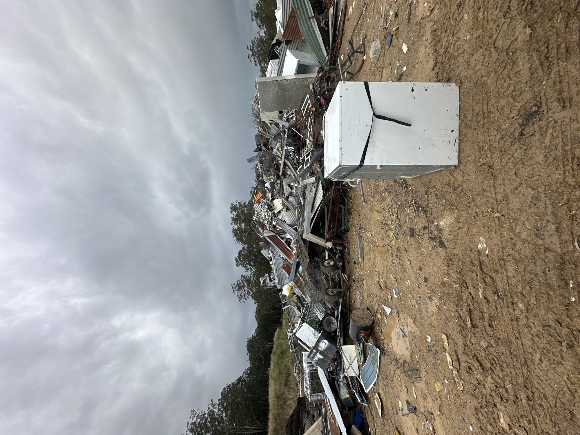 The image depicts a large pile of scrap metal and discarded appliances in an outdoor junkyard. The foreground is dominated by muddy ground with tire tracks, suggesting recent vehicle activity. A white appliance, possibly a washing machine or dryer, stands prominently in the mid-ground, secured with black straps. Behind it, a vast heap of mangled metal, corrugated iron sheets, and various machinery parts forms a chaotic landscape stretching towards a tree line under a cloudy, overcast sky. The scene is devoid of people and any discernible signs of urban infrastructure, placing it in a rural or industrial outskirts setting. The specific location in Raleigh, Australia, is not visually indicated by any landmarks or signage within the frame.