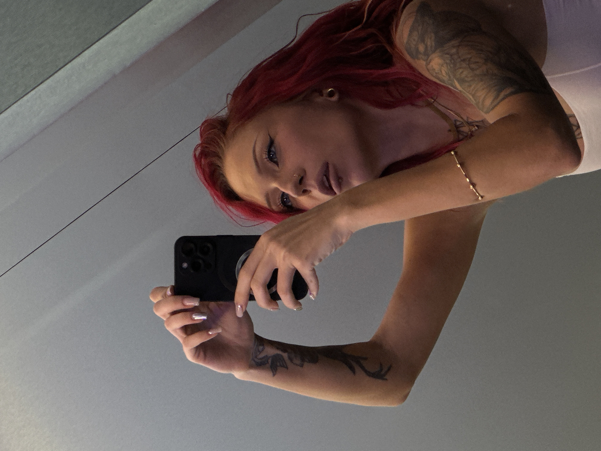 A person with red hair, tattoos on their arms and upper chest, and a nose piercing is taking a selfie with a black smartphone. The background appears to be a light gray surface, possibly a wall or ceiling. The person is wearing a white top and a gold bracelet on their arm. There is no discernible evidence indicating the location to be Kuta, Indonesia.