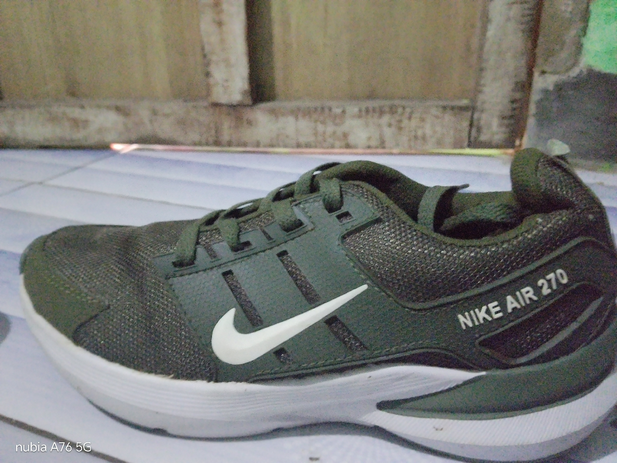 The image shows a close-up, side view of a dark green Nike Air 270 sneaker placed on a tiled floor. The sneaker is the primary subject, with the Nike swoosh logo visible in white on the side. Text that reads "NIKE AIR 270" is also visible on the heel of the shoe. The background consists of a wall with a textured, possibly concrete or wood paneling, and a lighter-colored tiled floor. The lighting appears to be artificial and slightly dim, casting soft shadows. The timestamp "nubia A76 5G" is visible in the bottom left corner. The image was taken in Binuang, Indonesia. There are no people or any observable actions in the image.