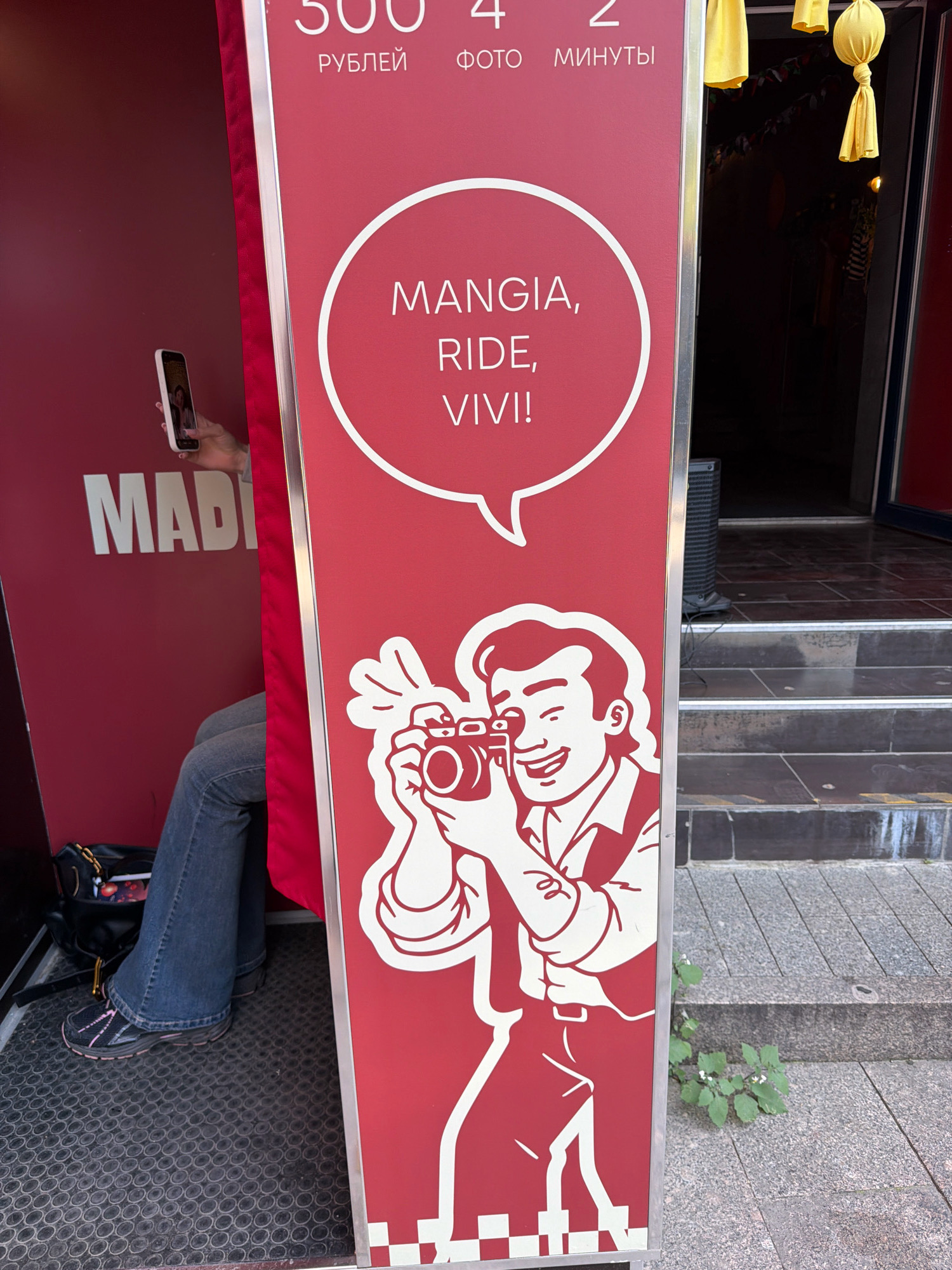 The image depicts a red photo booth with a white outline drawing of a man holding a camera. Text on the booth in white letters reads "MANGIA, RIDE, VIVI!". Above this, in smaller white lettering, are the Russian words "РУБЛЕЙ", "ФОТО", and "МИНУТЫ" which translate to "RUBLES", "PHOTO", and "MINUTES" respectively. The number "500" is visible to the left of "РУБЛЕЙ", and "4" is visible to the left of "ФОТО". A person is seated inside the booth, wearing blue jeans and a black and pink athletic shoe. They are holding a white smartphone up, as if taking a photo or video. A black backpack rests on the floor of the booth next to their feet. The booth appears to be situated outdoors on a tiled walkway with steps leading up to a dark entrance. Yellow fabric decorations hang from above. The scene is located in Moscow, Russia, as indicated by the Russian text on the photo booth.