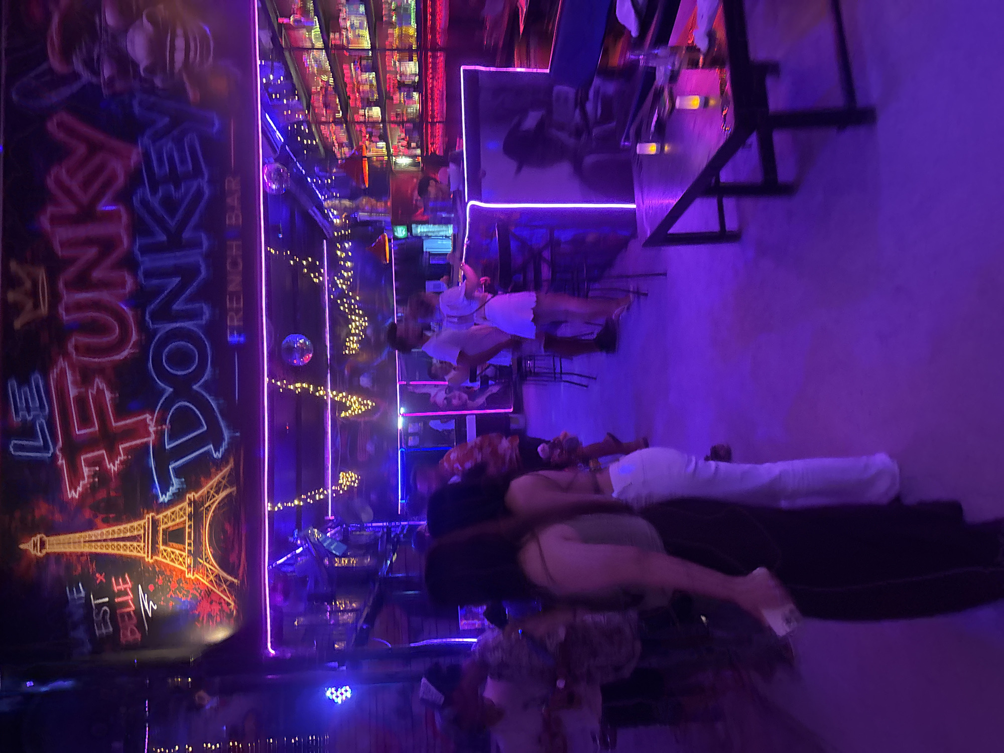 The image depicts the interior of a bar named "Le Funky Donkey - French Bar" in Đà Nẵng, Vietnam. The bar is dimly lit with vibrant neon purple and pink lighting, creating a lively atmosphere. A prominent graffiti-style mural of the Eiffel Tower with the text "La vie est belle" (Life is beautiful) is visible on the left side of the wall. The bar's name, "Le Funky Donkey," is displayed in large, neon letters, accompanied by a drawing of a donkey wearing sunglasses. Behind the bar, shelves are stocked with various bottles and glassware, illuminated by pink LED lights. Several people are present, including patrons standing and mingling, and staff behind the bar. The scene appears to be during evening hours, suggesting a social gathering or entertainment venue.