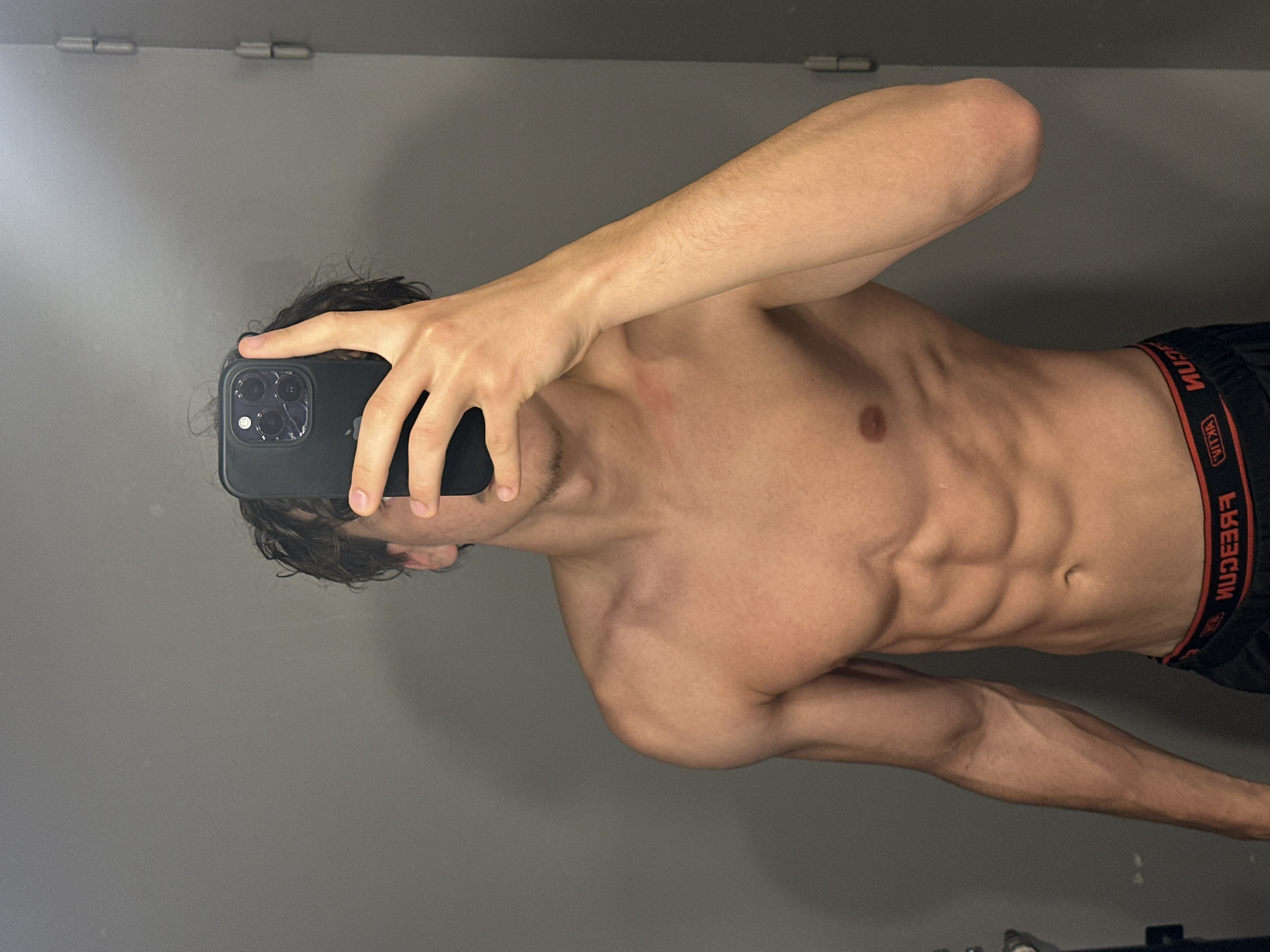 A shirtless young man stands in a dimly lit room, holding a smartphone to take a mirror selfie. He is facing the camera, with his left arm raised to obscure his face. His abdomen is visible, showing well-defined abdominal muscles. He is wearing black boxer briefs with a red and white band. The background appears to be a plain, grey wall with a metal door hinge visible on the right side of the frame. The lighting is dim and casts shadows, creating a moody atmosphere. The specific location in Anduze, France cannot be determined from the image.