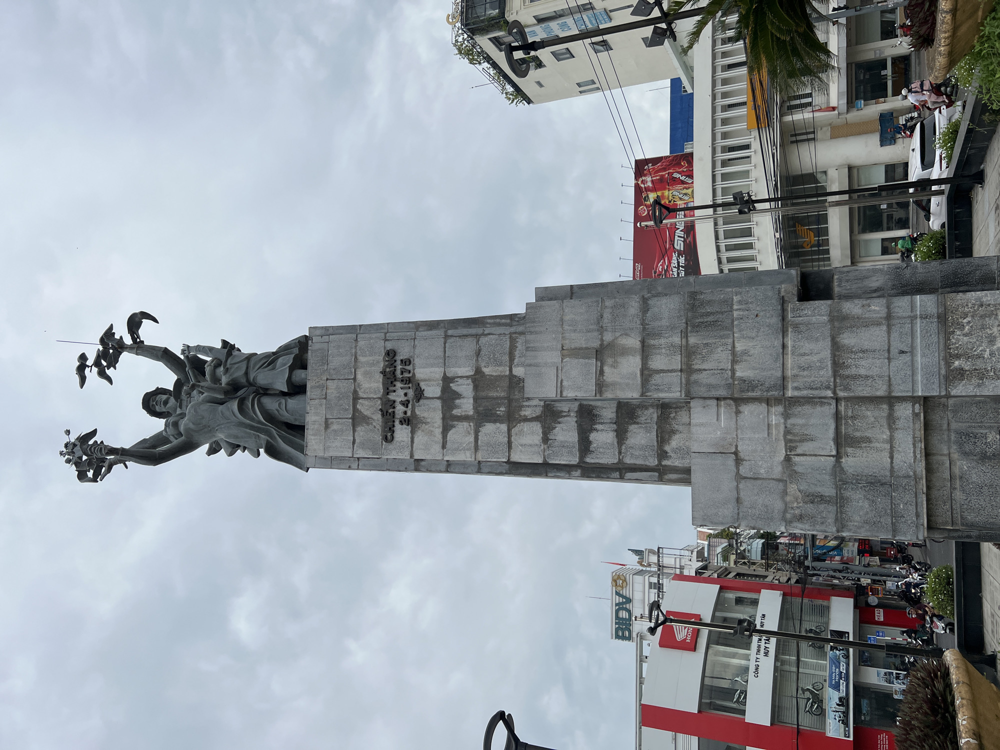 The image shows a large stone monument with a statue at its peak. The statue depicts several figures raising their arms, holding what appear to be birds or doves. The monument is inscribed with the words "CHIEN THANG 2-4-1975". In the background, a city street is visible, with buildings, traffic, and people. A Honda dealership with red and white signage is prominent on the left. Other buildings have various advertisements and signs. The sky is overcast. This monument is located in the city of Nha Trang, Vietnam, and it is known as the Victory Monument, commemorating the liberation of Nha Trang on April 2, 1975.