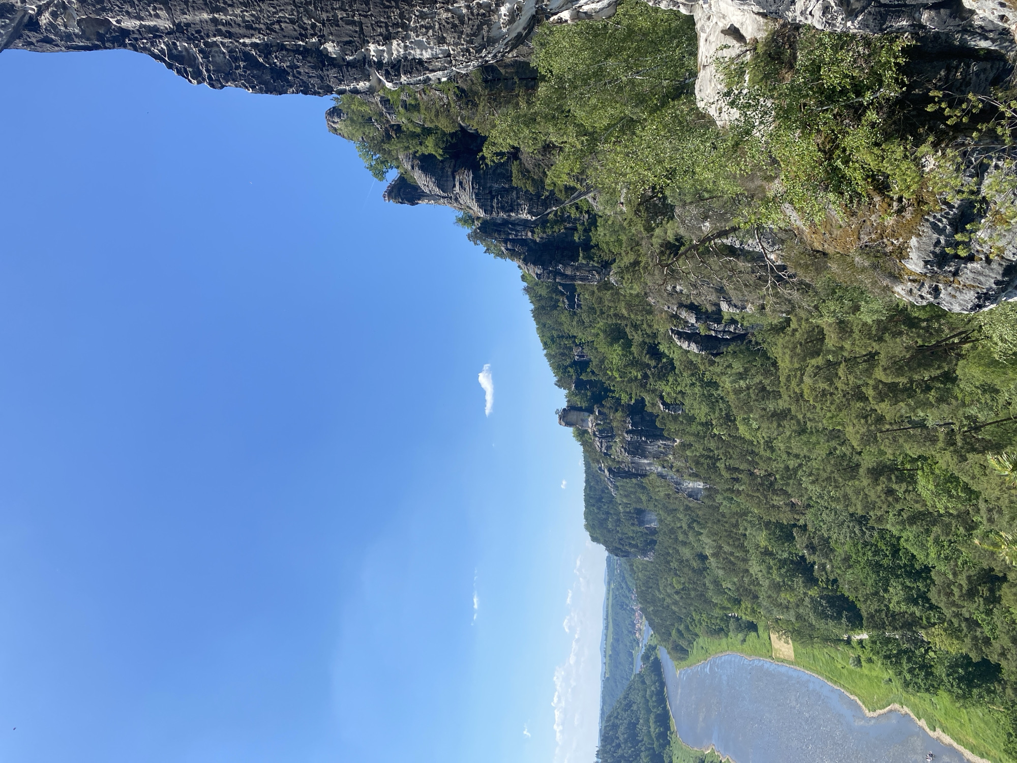 The image depicts a vertical panorama of a landscape in Lohmen, Germany. The scene features a geological formation, likely part of the Elbe Sandstone Mountains, with jagged cliffs covered in dense green trees. Below the cliffs, a river or body of water is visible, with greenery lining its banks. The sky above is a clear, bright blue with only a few sparse clouds. There are no people visible in the image.
