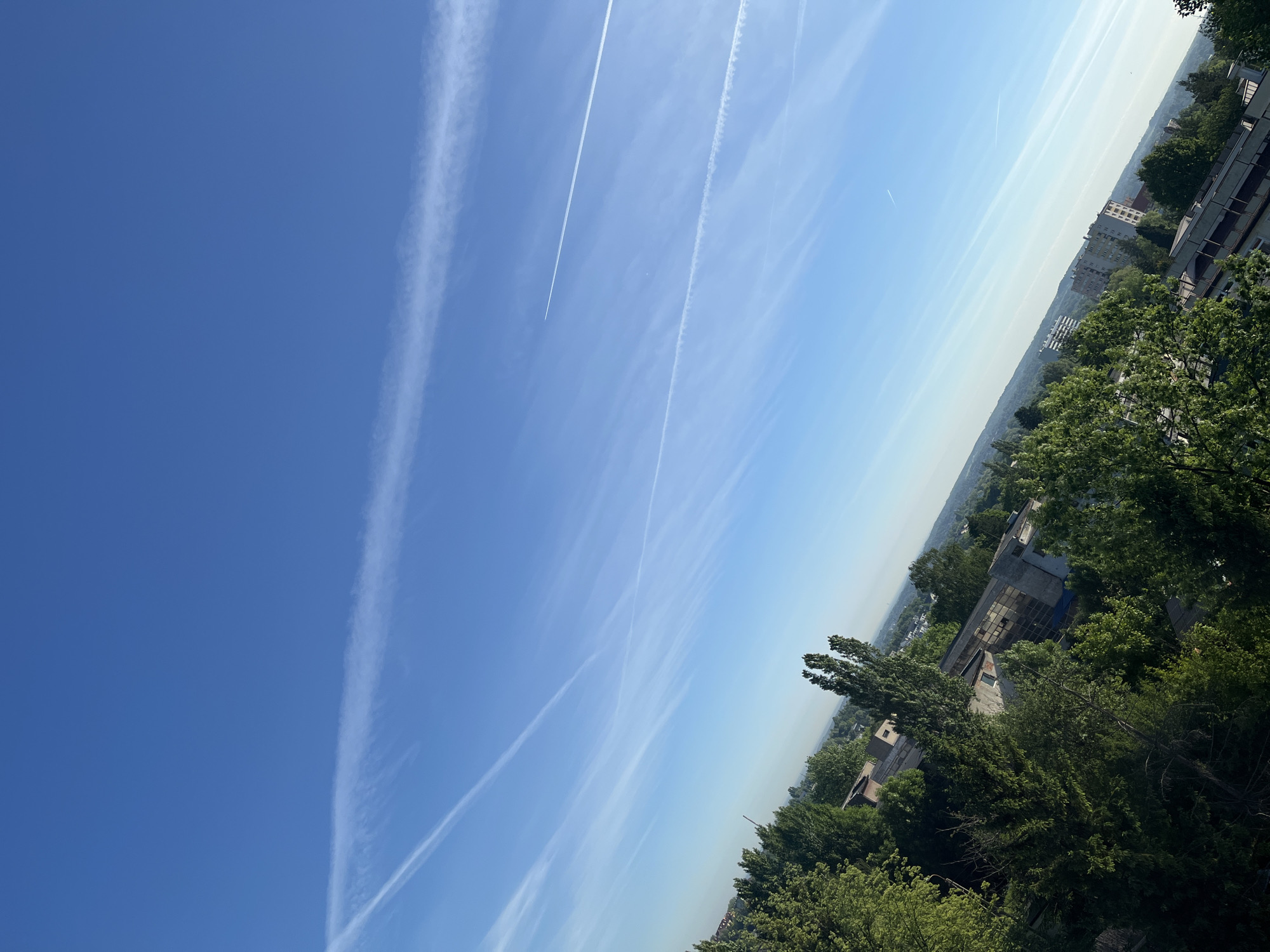 The image presents a cityscape under a blue sky with contrails. Buildings and trees are visible in the lower right of the frame. The sky transitions from a deep blue on the left to a lighter shade on the right, where a layer of haze obscures the horizon. Several streaks of contrails cross the sky. The photo appears to have been taken from an elevated position, offering a wide view of the city.