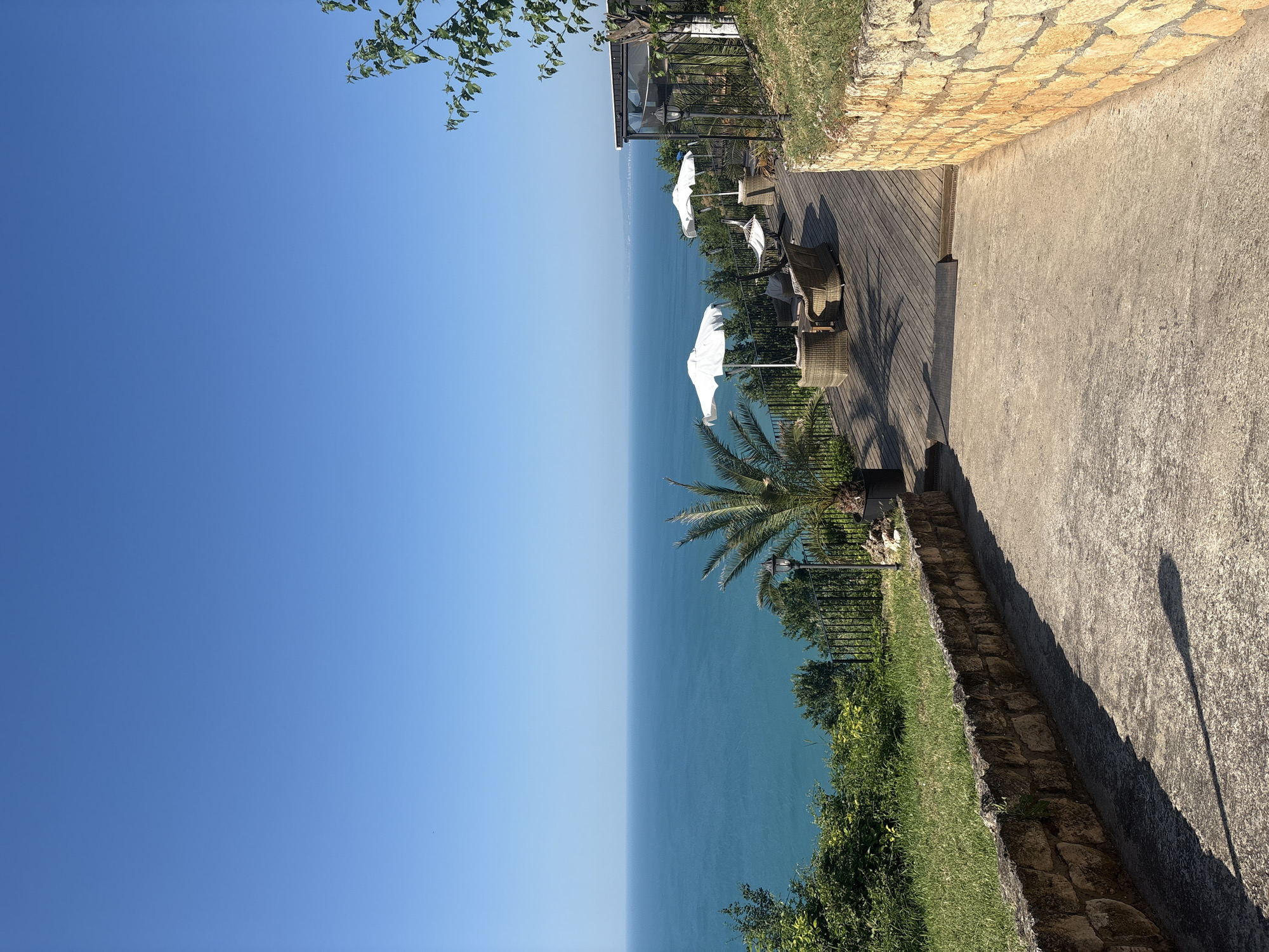 The image shows a bright, sunny day with a clear blue sky above a vast, turquoise ocean. In the foreground, a concrete pathway leads uphill, bordered by a rustic stone wall on the right and a grassy slope with a stone retaining wall on the left. Beyond the pathway, a wooden deck overlooks the ocean. The deck is furnished with wicker chairs, tables, and two white umbrellas, suggesting an outdoor seating area. A palm tree stands to the left of the deck. Further back, lush green foliage and trees descend towards the ocean. A black metal fence separates the deck area from the grassy slope. In the distance, a hazy coastline is visible. The overall scene depicts a serene coastal setting, possibly a restaurant or resort with a view. There are no people visible in the image.