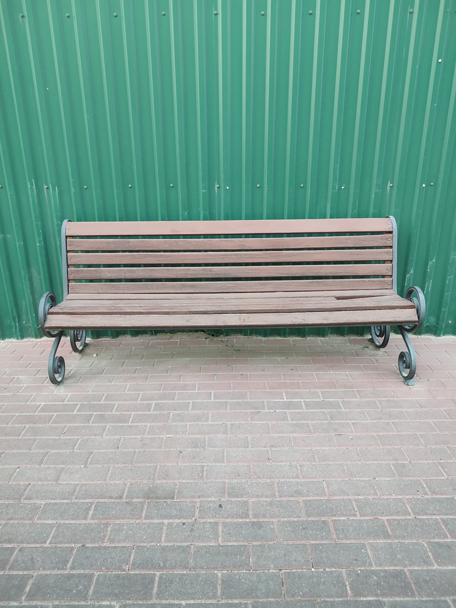 A wooden bench with metal scrollwork legs sits on a brick sidewalk in front of a green corrugated metal fence. The bench is empty. The scene appears to be outdoors in an urban environment.