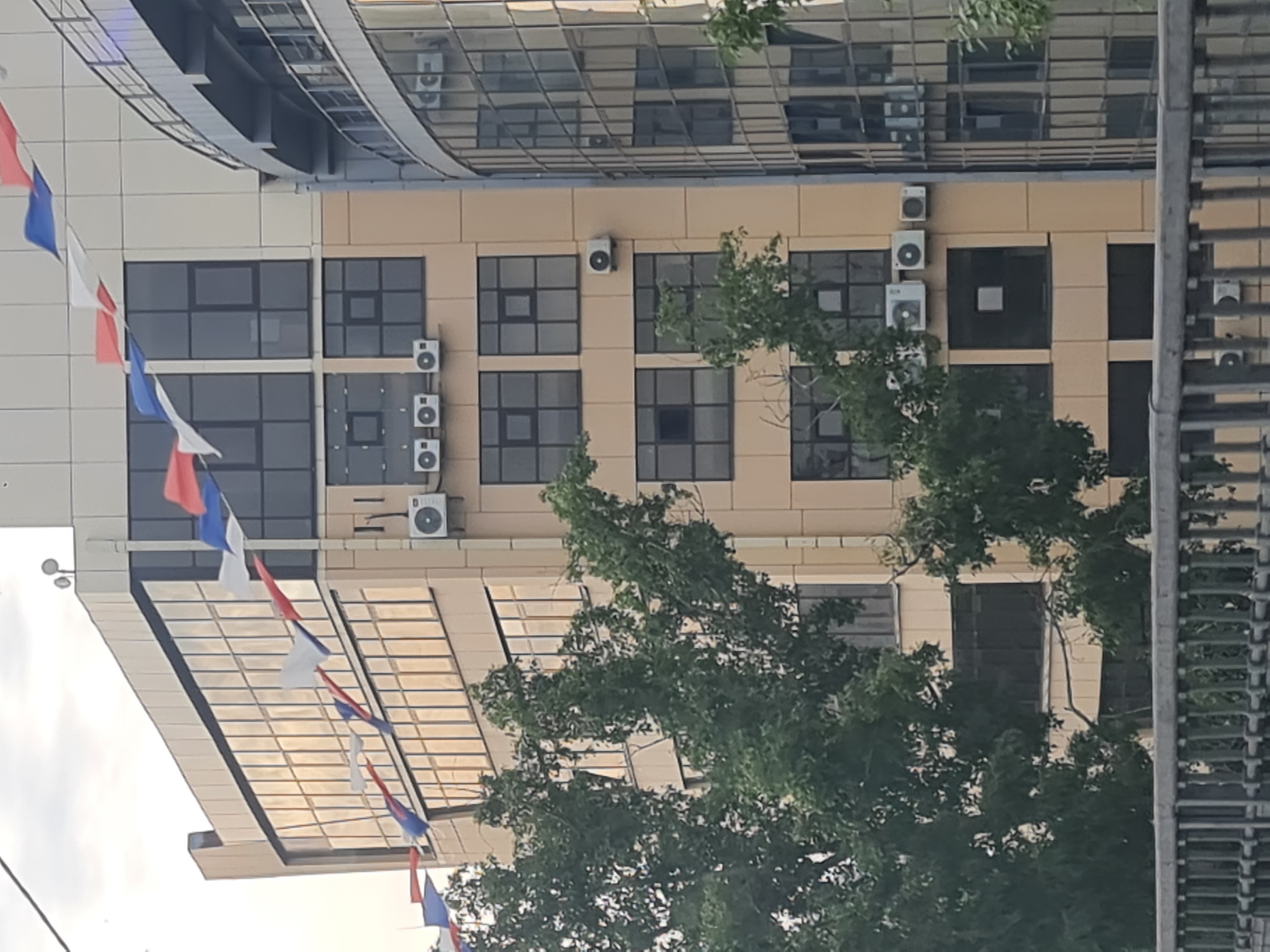 The image shows a building facade in Irkutsk, Russia. The building has large windows with a reflective surface. There are several air conditioning units mounted on the exterior of the building. Decorative flags in red, white, and blue are strung across the top of the building. Trees with green foliage are in the foreground, partially obscuring the building. A metal fence is visible at the bottom of the image. The sky is overcast.