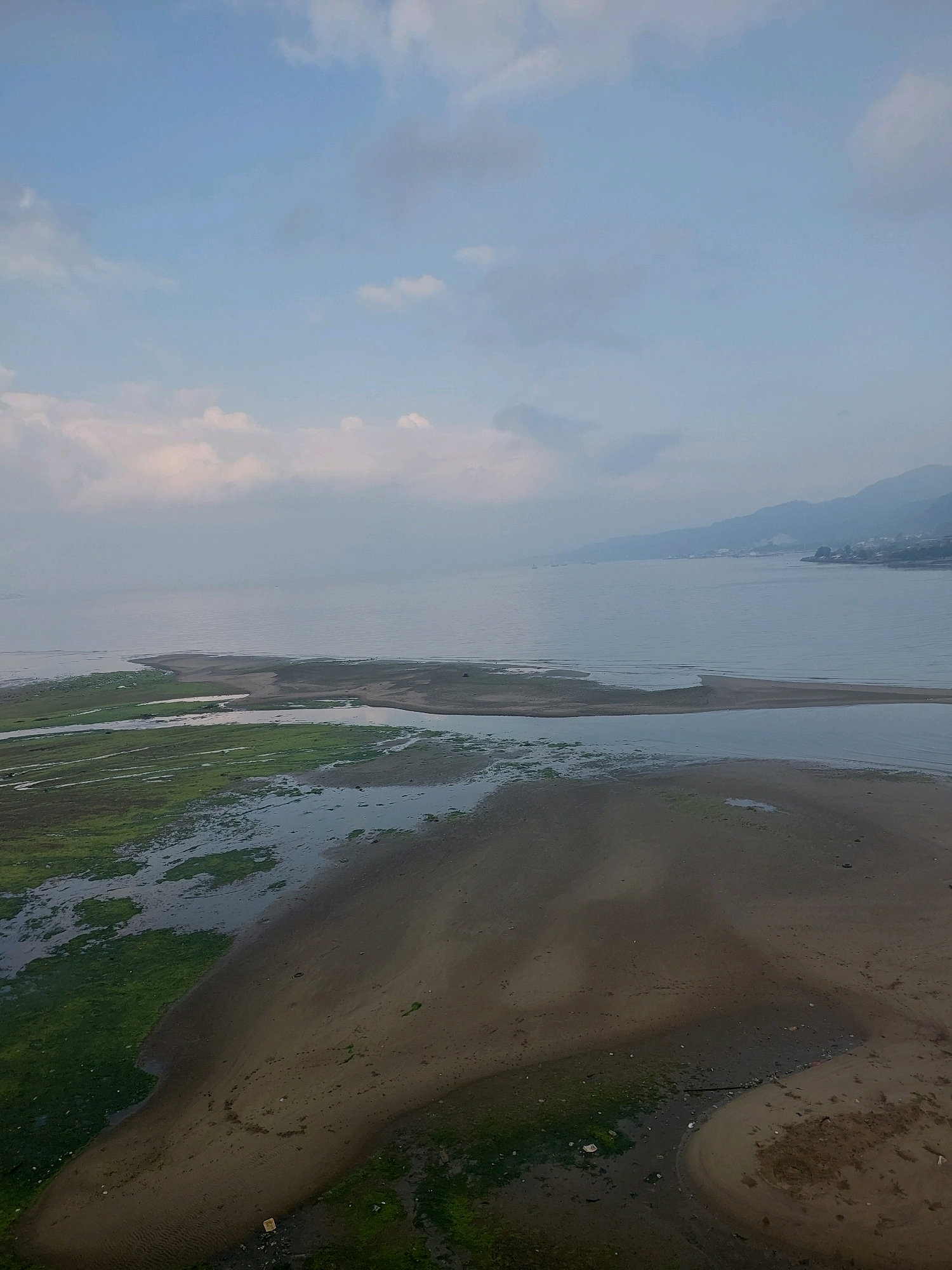 The image shows a coastal landscape with shallow water, sandbanks, and green algae-covered areas. In the foreground, there are sandy areas with ripples from water. Further back, there are patches of green algae and shallow pools of water. A sandbank extends into the calm body of water, which occupies the midground and background. The water stretches towards a hazy shoreline with hills and mountains in the distance. The sky is overcast with light clouds. The scene appears to be a natural, undeveloped coastal area. Given the visual elements of a coastal environment with hills and mountains meeting the sea, and the general visual characteristics which could be consistent with Southeast Asian geography, this scene could potentially be located in Ambon, Indonesia.