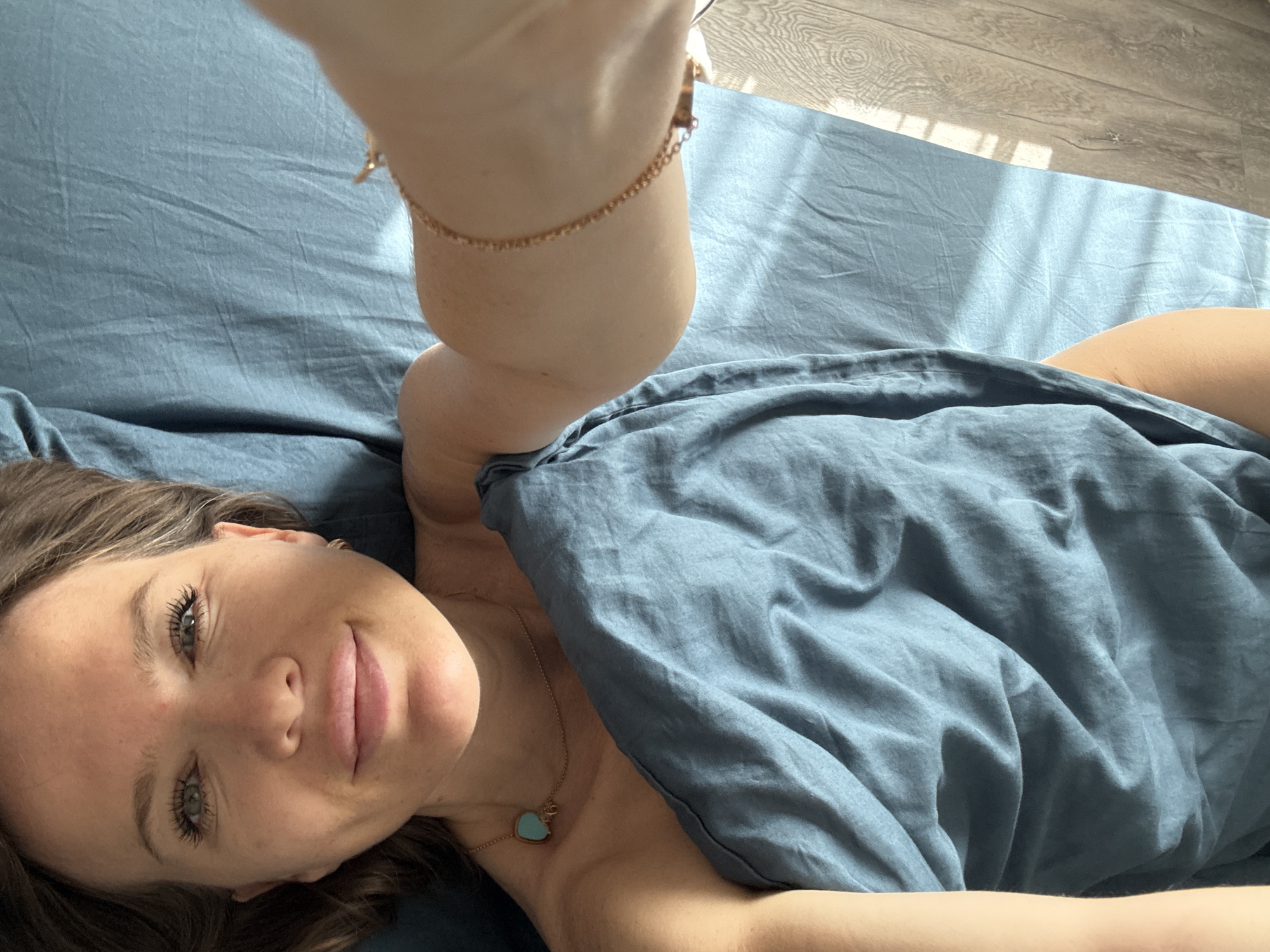 The image shows a young woman lying on a bed with blue sheets. She is looking up and smiling slightly at the camera. She has long, dark hair and is wearing a necklace with a blue heart pendant. Her arm is raised, and she is wearing a delicate gold bracelet on her wrist. The bed is situated on a wooden floor, and sunlight is streaming in through a window, casting striped shadows on the sheets and floor. The lighting suggests it is daytime. The overall mood is relaxed and intimate. The location is identified as Khimki, Russia, but there are no specific visual cues in the image to confirm this.