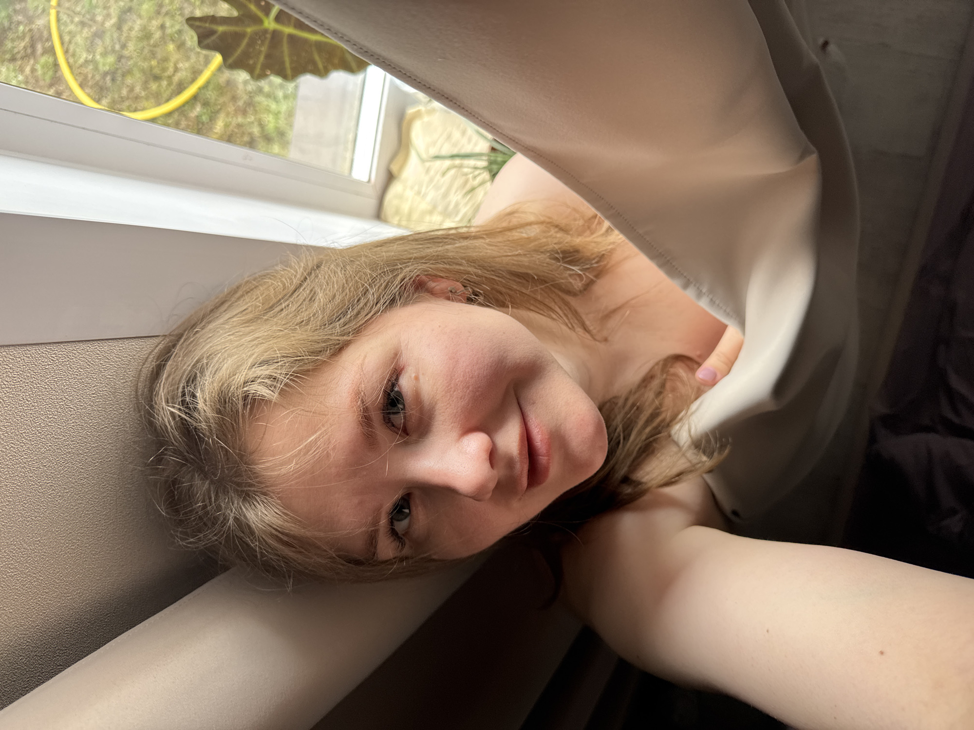A light-skinned person with light brown hair is holding a camera and taking a selfie. The person is positioned near a window. Through the window, blurred foliage and a yellow garden hose are visible. The person is partially obscured by a light-colored curtain or sheet. A dark object or space, possibly a bed or dark furniture, is in the background on the right side of the frame. The location is undetermined, and there is no information in the image to connect it to Gornyi (Dvurechenskoe S/P), Russia.