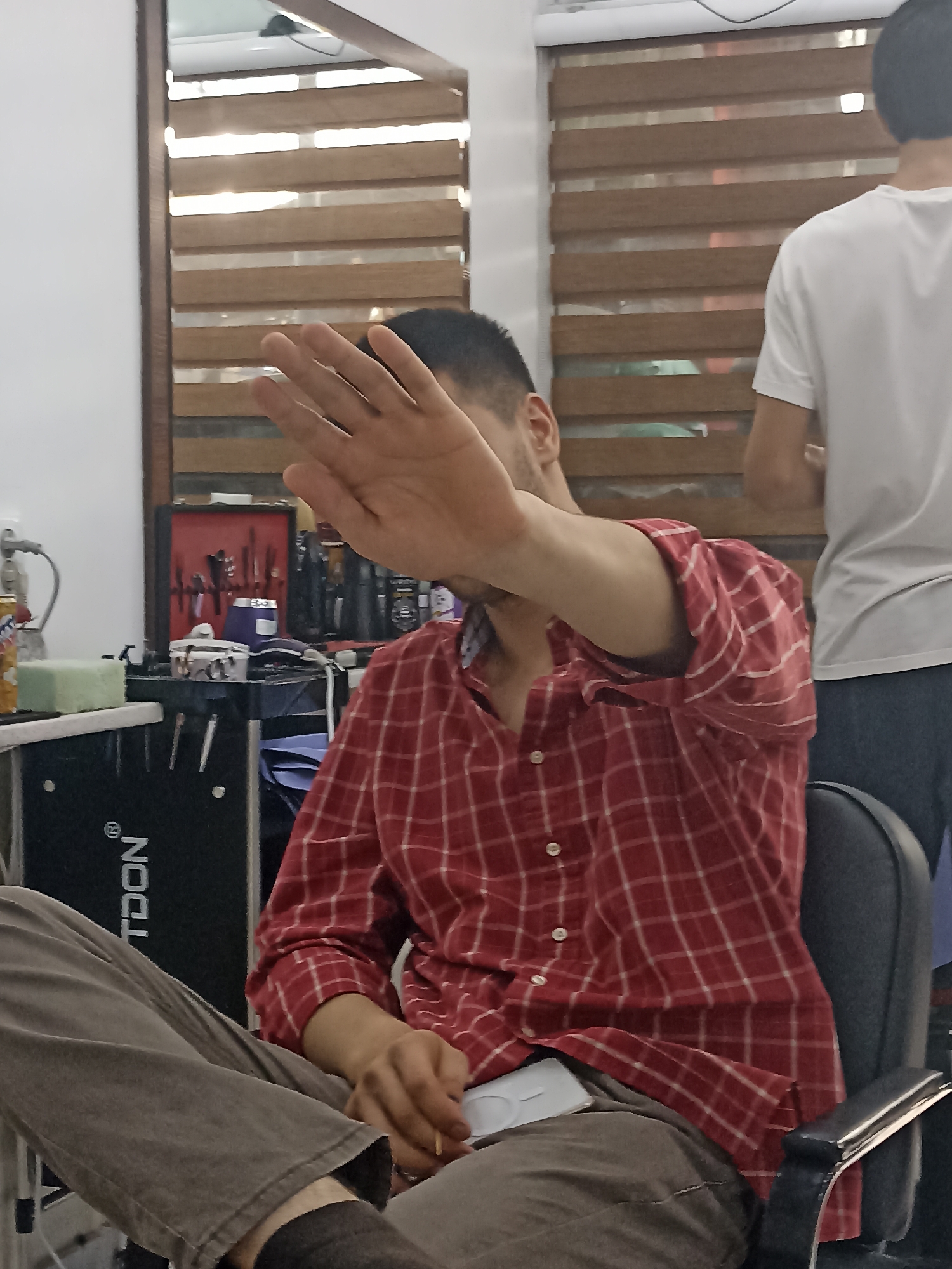 A young man with short, dark hair sits in a barber's chair, holding his right hand up to shield his face. He is wearing a red and white plaid shirt and grey pants. A white cell phone is visible on his lap. In the background, a barber's station is visible with various tools, and another person in a white t-shirt stands behind him. The environment appears to be a barbershop, likely in Dushanbe, Tajikistan, as indicated by the presence of a "TDON" branded piece of equipment, which is a Tajik company. The room has wooden blinds on a window.