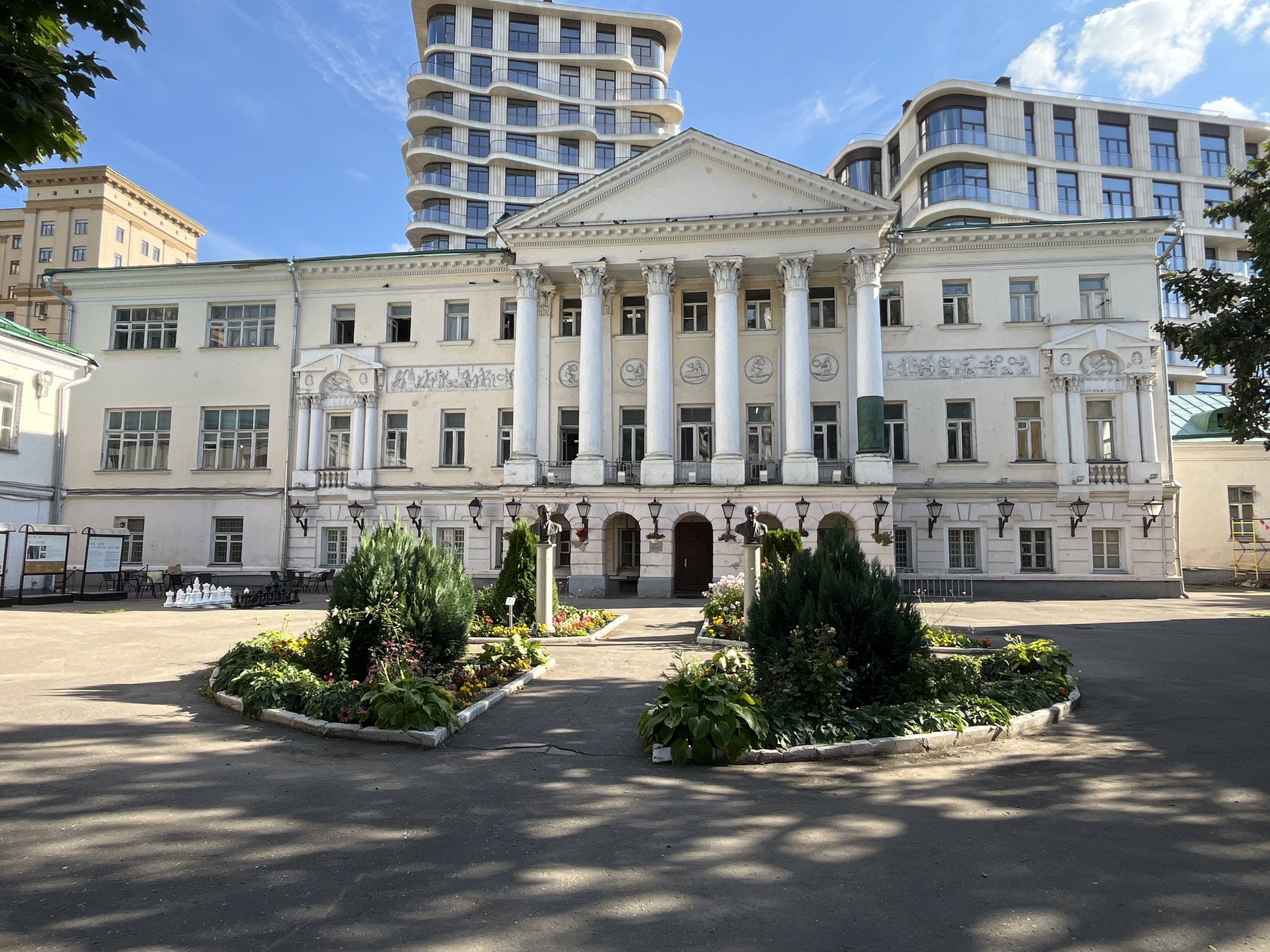The image shows the facade of a large, neoclassical building with a prominent portico featuring eight white Ionic columns. The building is pale yellow and has many windows. Behind and to the right of this building, a modern, multi-story building with a curved facade and many balconies is visible. In front of the neoclassical building, there are manicured flower beds with various green plants and colorful flowers. Two stone busts are placed on pedestals within the garden area, facing the entrance. A paved courtyard with shadows cast by trees extends in front of the garden. To the left, tables and chairs are set up, and a large outdoor chessboard with white and black pieces is arranged on the ground. Further to the left, informational boards are mounted on stands. The sky is blue with scattered clouds. The scene appears to be a public space or educational institution. This location is the Russian State University for the Humanities (RGGU) on Miusskaya Square in Moscow, Russia.