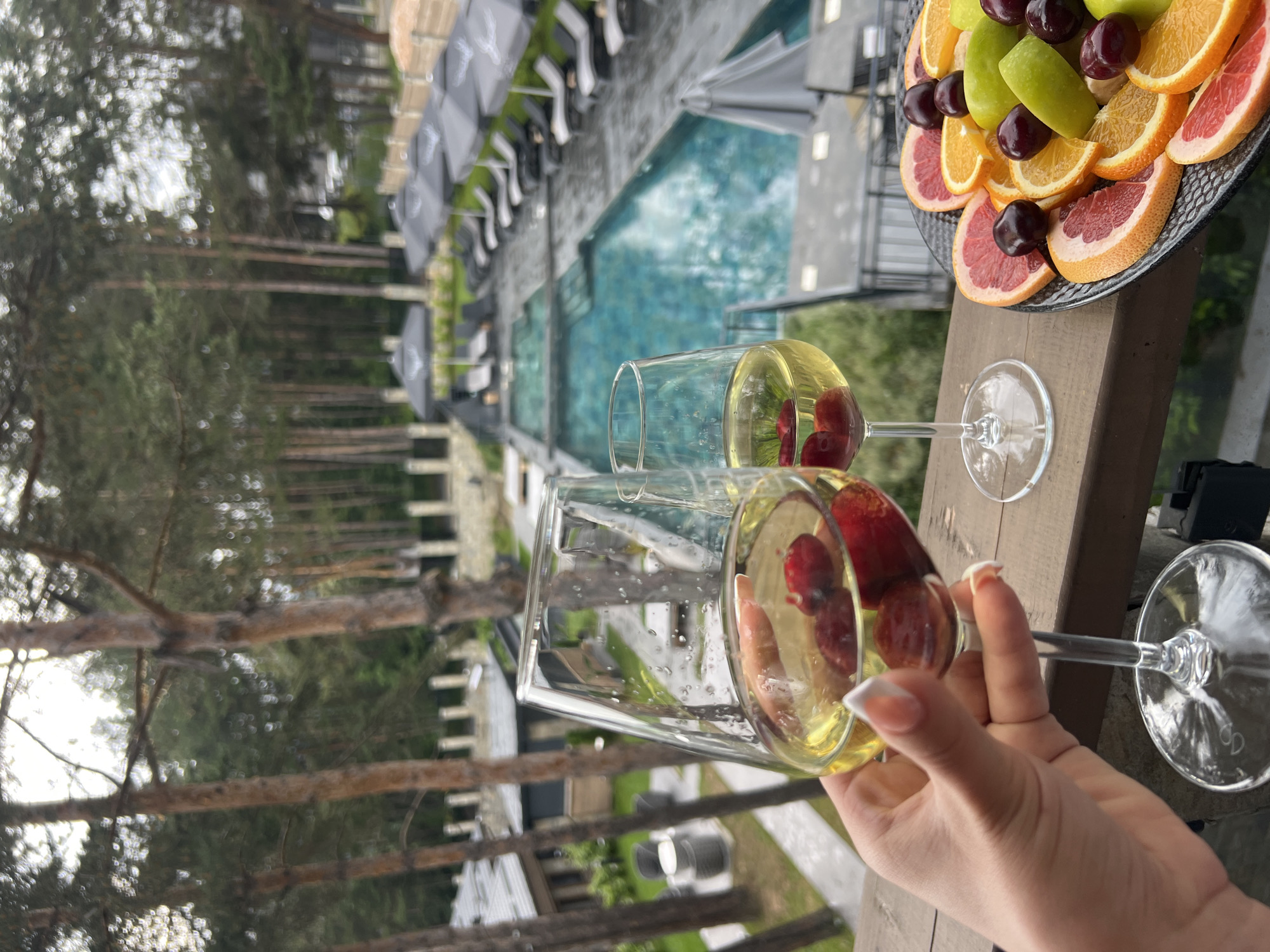 A hand with a French manicure holds a champagne glass filled with a yellow beverage and two red cherries. Another champagne glass, also with cherries, is visible in the background. A platter of sliced fruits, including oranges, grapefruit, apples, and cherries, sits on a wooden surface. Behind the glasses and platter, a swimming pool with blue water is surrounded by lounge chairs and umbrellas. Pine trees and a building are visible in the distance. The scene appears to be outdoors, possibly on a balcony or patio, with a relaxed and leisurely atmosphere. The lighting suggests it could be daytime.