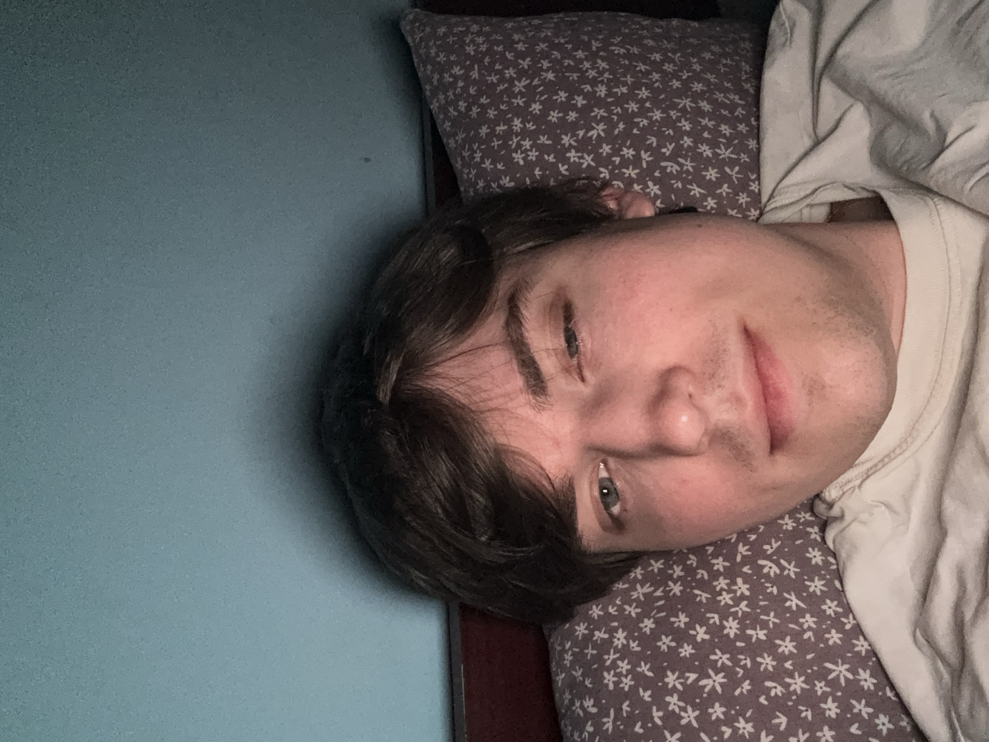 The image is a close-up selfie of a young Caucasian male lying in bed. He has light brown hair and blue-green eyes, and is wearing a light-colored t-shirt. The background consists of a teal-colored wall and a pillow with a white floral pattern on a dark gray background. The lighting suggests it is nighttime. The image does not contain information that would allow for the determination of its location in the city of Dnipro, Ukraine.