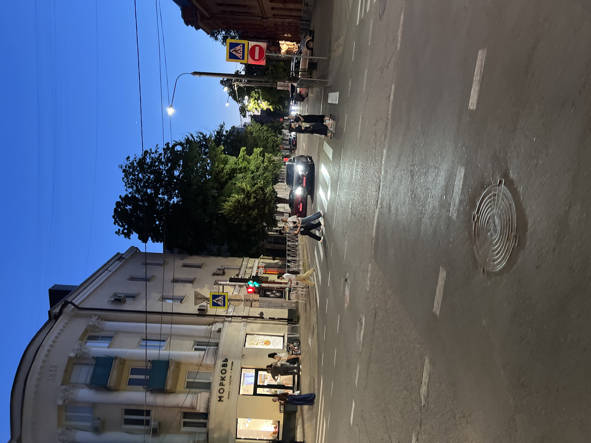The image depicts a street scene in Krasnodar, Russia, at dusk or early evening. The sky is a deep blue, indicating the transition from day to night. The wet asphalt suggests recent rain or a damp atmosphere. The scene is illuminated by streetlights and the headlights of vehicles.
Several people are visible. Two individuals are crossing a pedestrian walkway in the middle of the street, seemingly engaged in conversation. Further down the street, more people are walking on the sidewalk near a brightly lit storefront. One person is seen standing near the entrance of the store, and another is walking away from it.
Objects in the scene include cars, traffic lights (showing green and red lights), road markings (white stripes and a pedestrian crossing), and a manhole cover. A building with multiple windows and architectural details lines one side of the street. Trees with lush green foliage are also present, adding to the urban landscape.
There are a few signs visible. One sign is a yellow diamond with a blue pedestrian icon, indicating a pedestrian crossing. Another sign above it is red and circular with a white horizontal bar, which is a "no entry" or "closed" sign. The building facade has visible text, including "МОРКОВЬ" which translates to "CARROT" in Russian, suggesting it might be a shop or cafe. The text below it appears to be "СОКИ • СМУЗИ • ЯГОДЫ", meaning "JUICES • SMOOTHIES • BERRIES".
The overall atmosphere is that of a normal evening in a city, with people going about their activities under the evening sky. The wet street and subdued lighting create a slightly muted, tranquil mood.