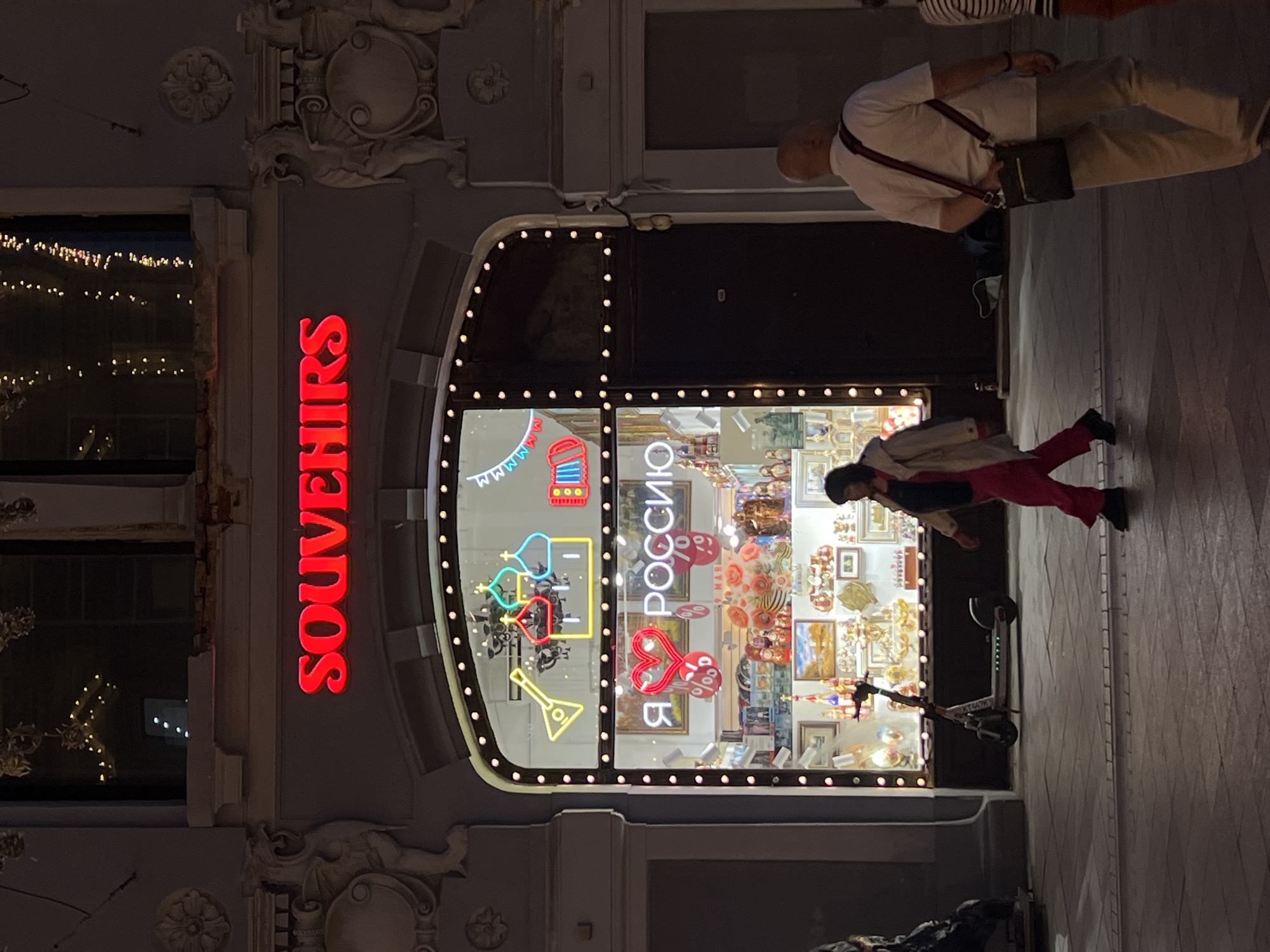 The photograph shows the exterior of a souvenir shop in Moscow, Russia, at night. The shop's facade features a prominent, vertical red "SOUVENIRS" sign. The shop window display includes neon signs shaped like Russian symbols such as matryoshka dolls, balalaikas, and onion domes, along with other traditional Russian souvenirs.
Several people are present. One individual is dressed in a white shirt and khaki pants, with a shoulder bag, is walking on the sidewalk. Another individual is standing near the shop window, wearing a coat or jacket with red pants. A scooter is parked near the shop entrance. The sidewalk appears to be paved with stone.