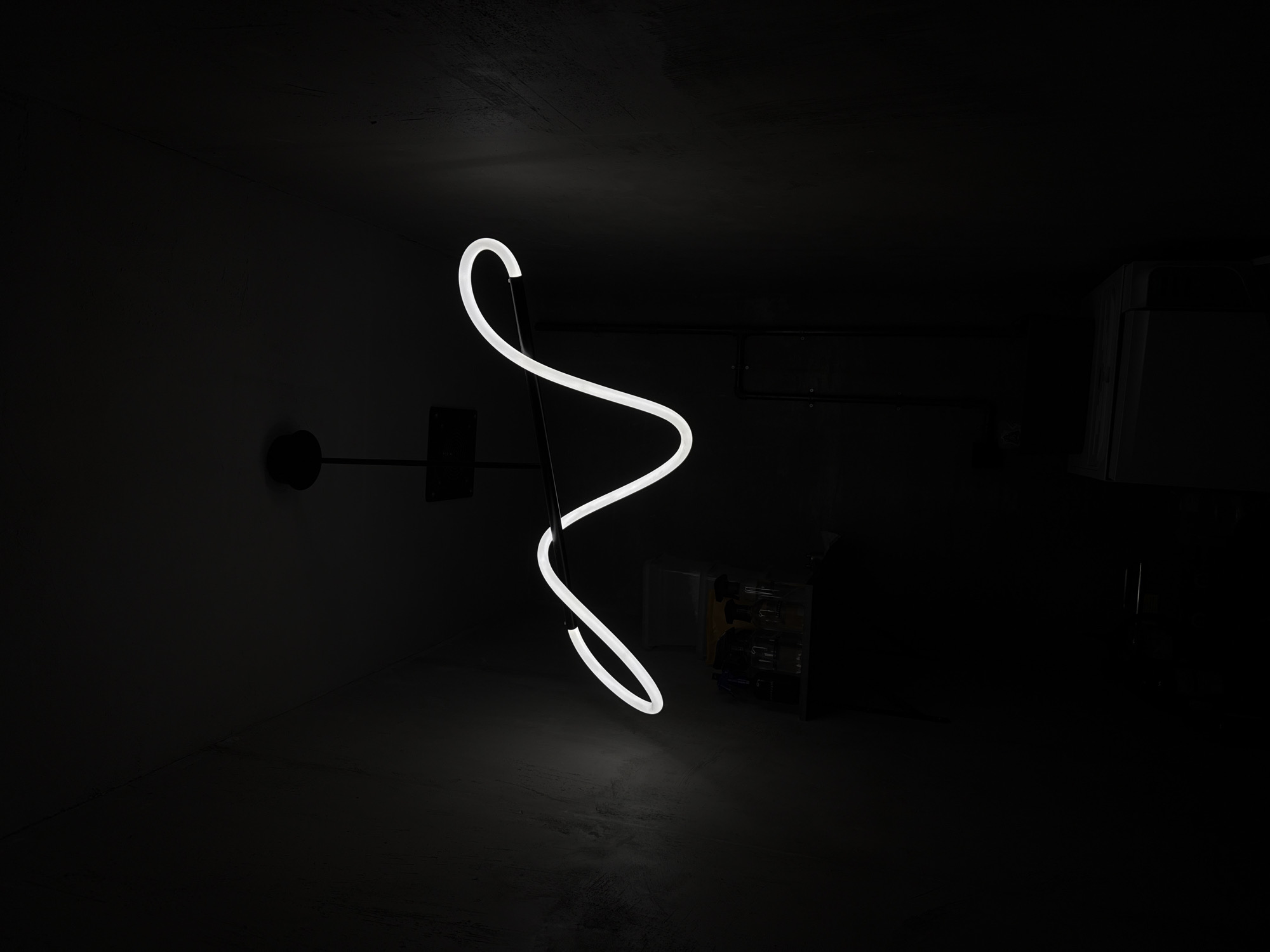 A dimly lit room features a striking, illuminated white neon sculpture. The sculpture consists of a winding, tubular form that appears to stand vertically from a dark base. The light from the sculpture is the primary source of illumination, casting a soft glow on the floor. The room itself appears to be a utilitarian space with dark walls and minimal furnishings. A black pole extends into the light. Other background objects are hard to discern in the darkness. Based on the details provided, it's not possible to confirm if the location is Moscow, Russia or to provide additional location details.