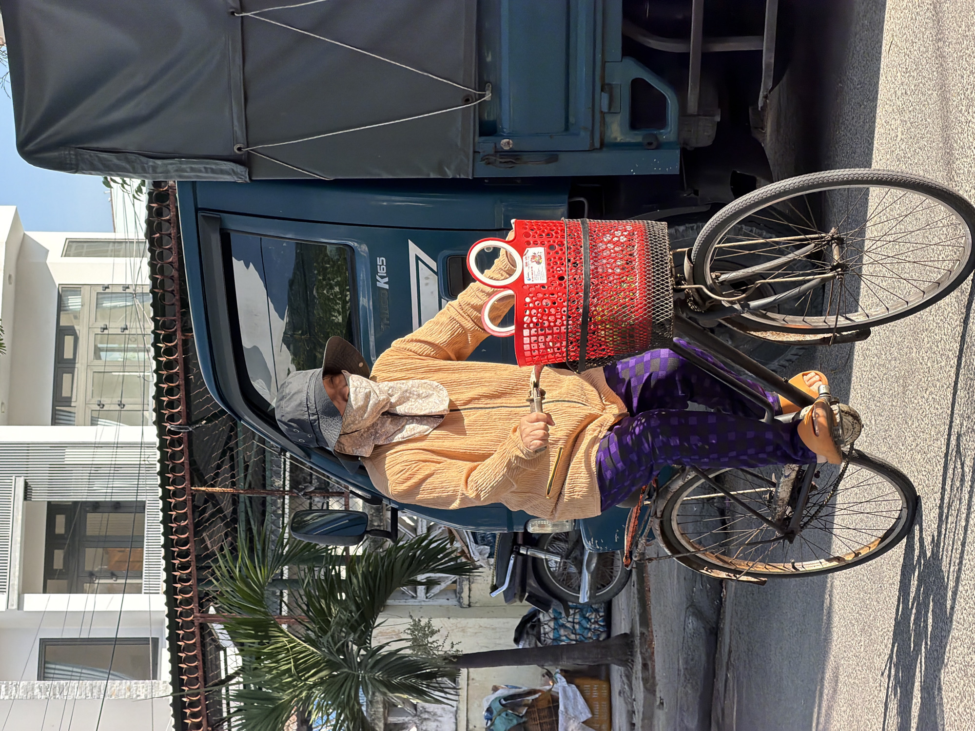 A woman wearing a hat and a face covering rides a bicycle down a street in Nha Trang, Vietnam. She is wearing an orange long-sleeved shirt and purple pants with a checkered pattern. Her bicycle has a red basket in front, which holds two round objects, possibly bottles or containers. A large blue truck is parked on the right side of the image, partially obscuring the woman and her bicycle. Behind them, a building with white and grey vertical siding is visible. A palm tree stands to the left of the woman, with a motorcycle parked behind it. The sun is casting shadows on the asphalt road, indicating it is daytime.