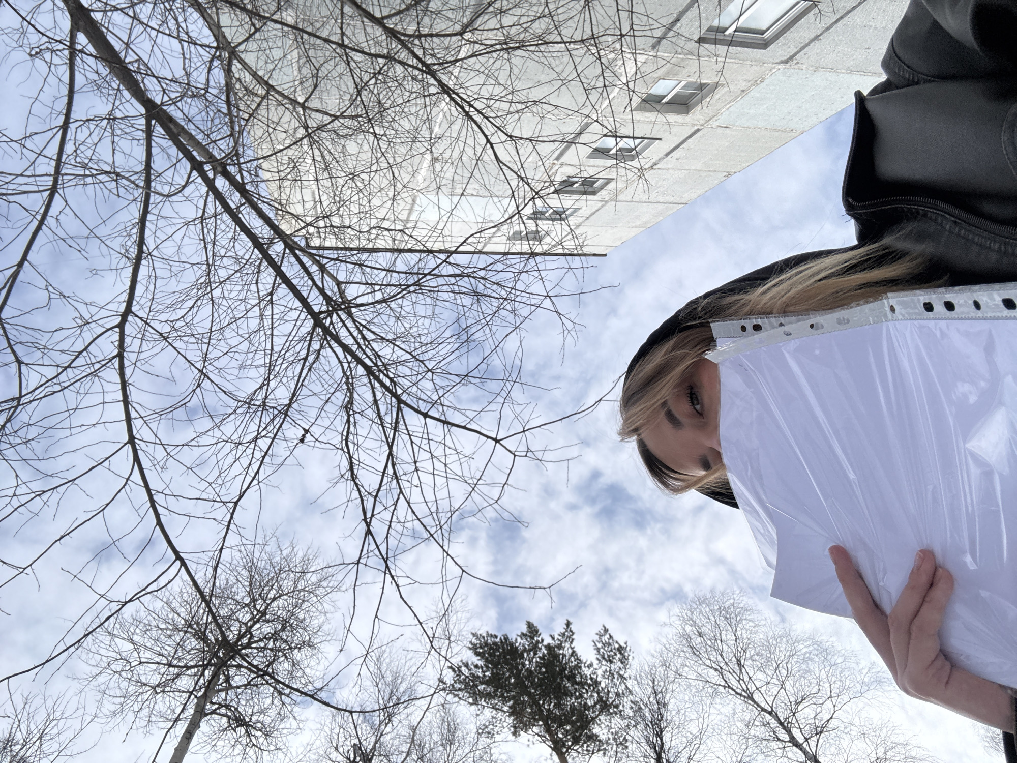 A low-angle shot depicts a blonde-haired person wearing a black hooded jacket holding a stack of papers in a plastic sleeve. The person is positioned in the right side of the frame. Behind the person, a pale blue sky streaked with white clouds is visible, framed by bare tree branches. A building with multiple windows is slightly out of focus and positioned upside down in the upper portion of the frame. The scene appears to be outdoors, likely in an urban area of Ust-Ilimsk, Russia.