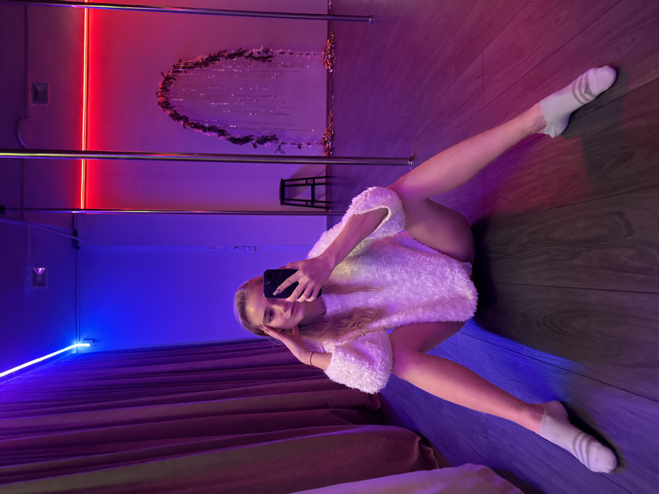 A woman in a fluffy white top and white socks takes a selfie in a room with vibrant red and blue neon lighting. A dance pole is visible in the background, along with a chair and a decorative garland. The room features wood flooring and a curtained area, creating a dynamic and modern aesthetic.