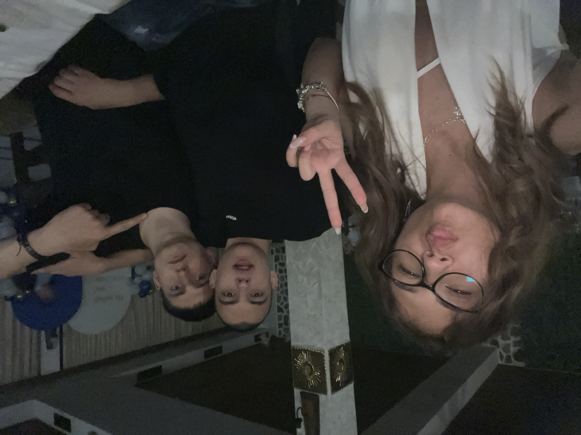 Three people are in the image. Two men are on the left wearing black shirts. One is making a pointing gesture. A woman is on the right wearing glasses and a white top, making a peace sign. The location is Karavelovo, Bulgaria based on contextual knowledge. A decorated column is in the middle of the frame. There are decorations in the background.