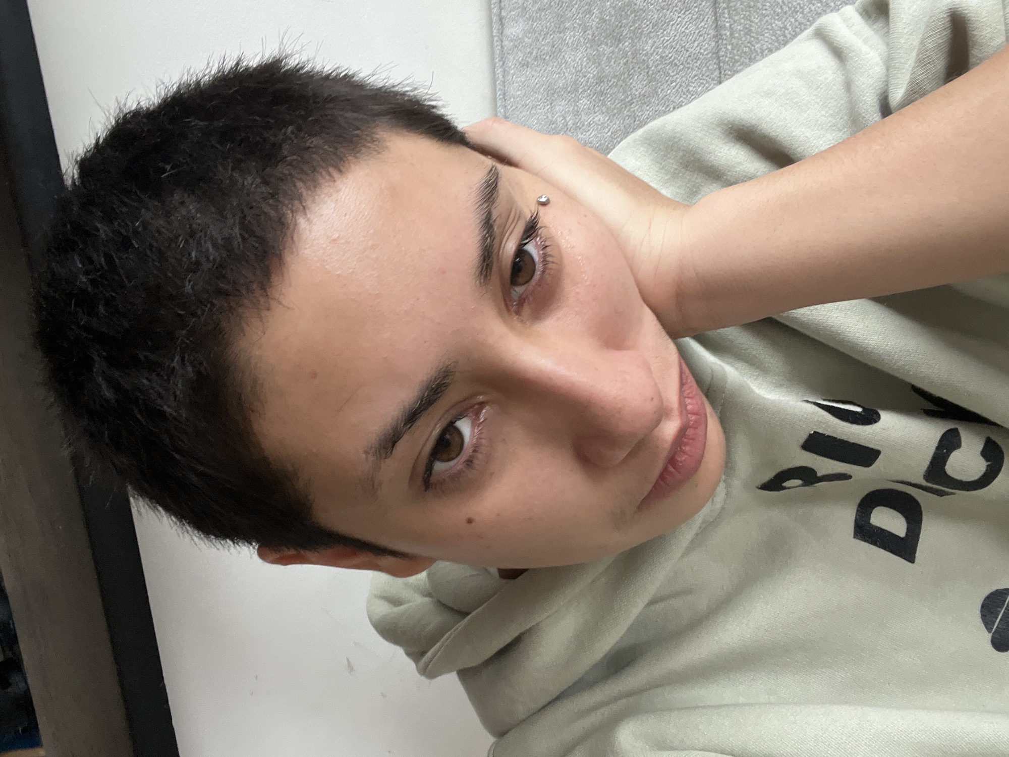 The image is a close-up selfie of a person with short dark hair, resting their head on their hand. They are wearing a light green hooded sweatshirt with black lettering on the front. A silver piercing is visible on their left cheekbone. The background is blurred but appears to be indoors, with a white wall and a gray surface that could be a couch. There are no identifiable landmarks or features that would indicate the location in Buenos Aires, Argentina.