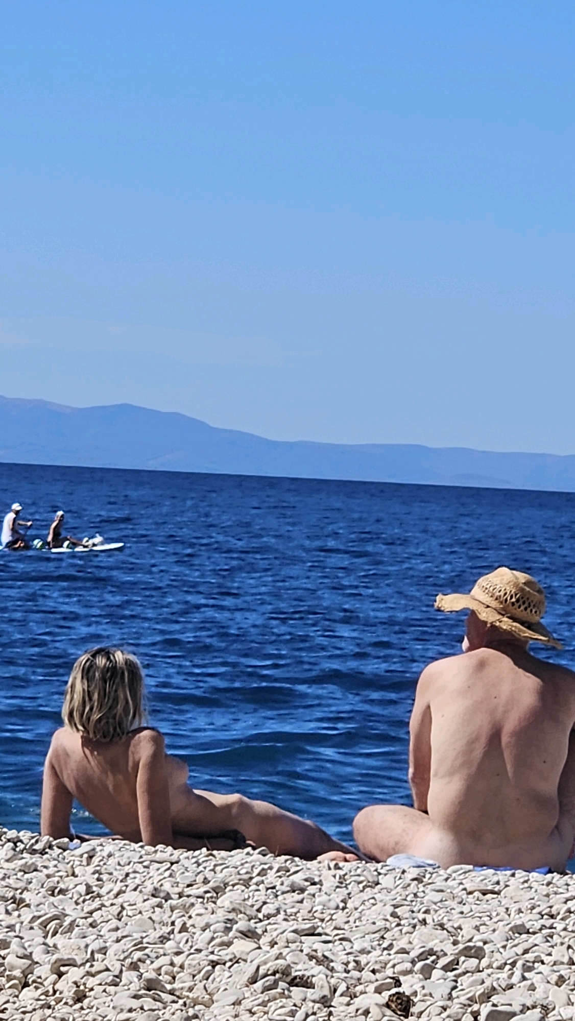 In Općina Podgora, Croatia, on a pebble beach, two individuals are present. In the foreground, a person with blonde hair is lying on their side, facing the sea, with their back to the camera. To their right and slightly behind, another person wearing a straw hat is seated with their back to the camera, also facing the sea. The sea is a deep blue, with gentle waves. In the distance, on the water, three figures are visible on a paddleboard. Further in the background, mountains form the horizon under a clear blue sky.