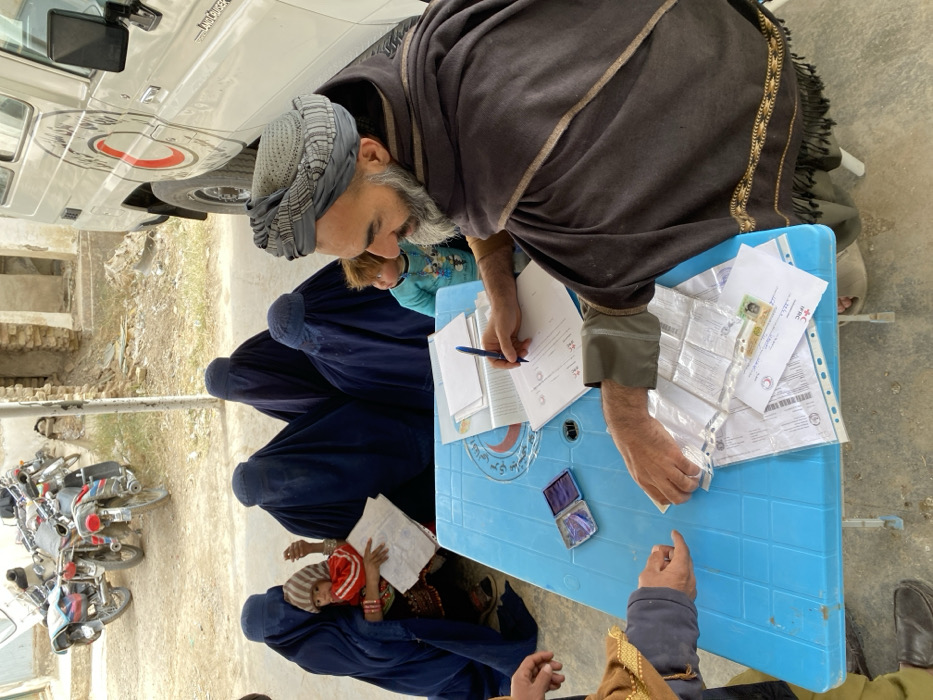 A man in a turban and shawl sits at a blue plastic table, reviewing documents and writing with a pen. Beside him are several women wearing full-body burqas, with a young child visible among them. Additional documents and a small box sit on the table, bearing the red cross emblem. Motorbikes are parked nearby, and a white vehicle with a red cross symbol is partially visible in the background.