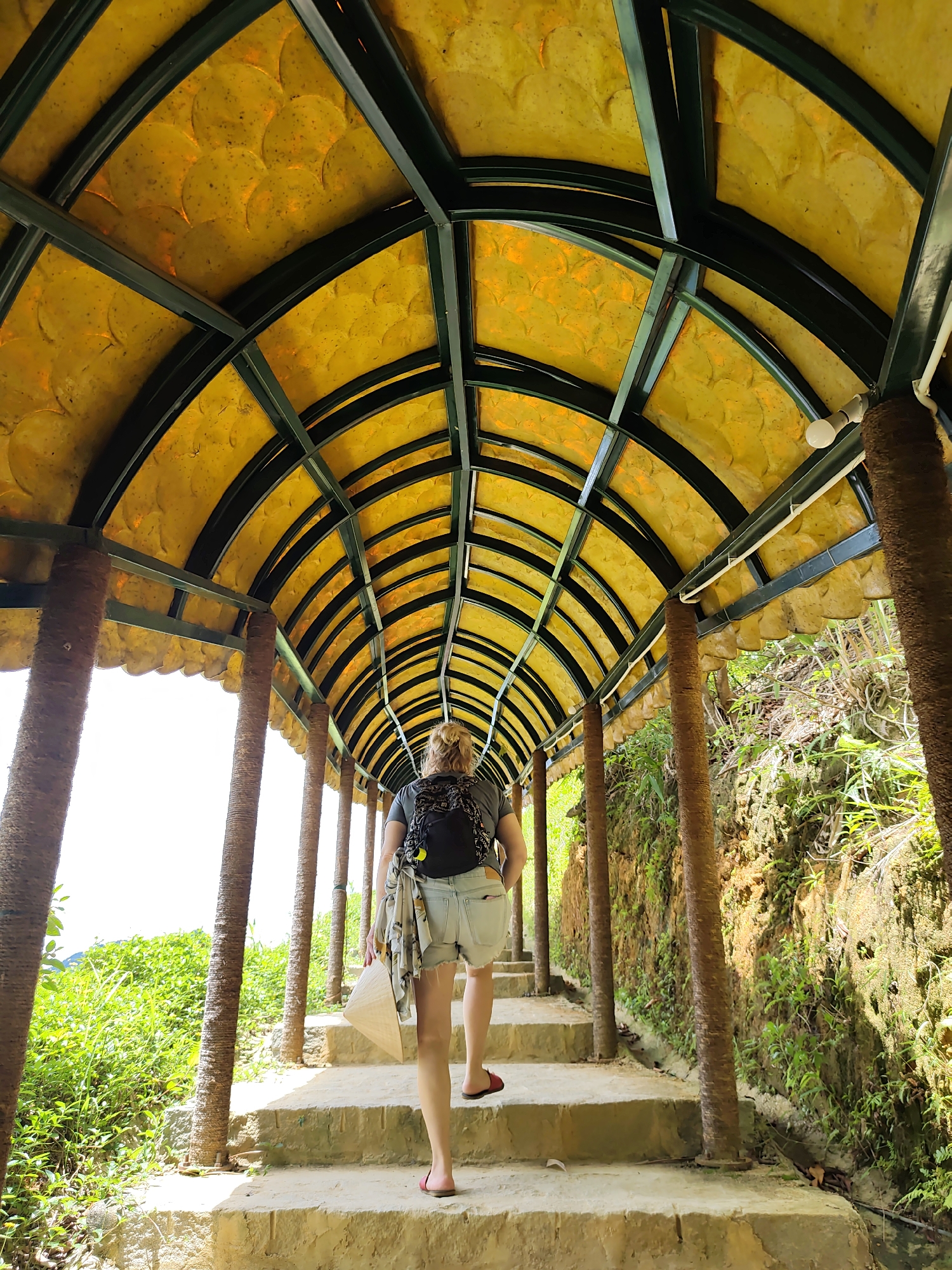 In an outdoor setting in Unknown, Unknown, a person walks up a stone staircase under a covered walkway. The walkway has a green metal frame and a yellow, textured ceiling. The walkway is supported by thick, textured columns. The person, facing away from the camera, wears a backpack, shorts, and a light-colored shirt draped over their shoulder. They are carrying a conical hat in their right hand. Lush green vegetation grows on the right side of the staircase, and similar greenery is visible to the left. The sky appears bright and possibly overcast.