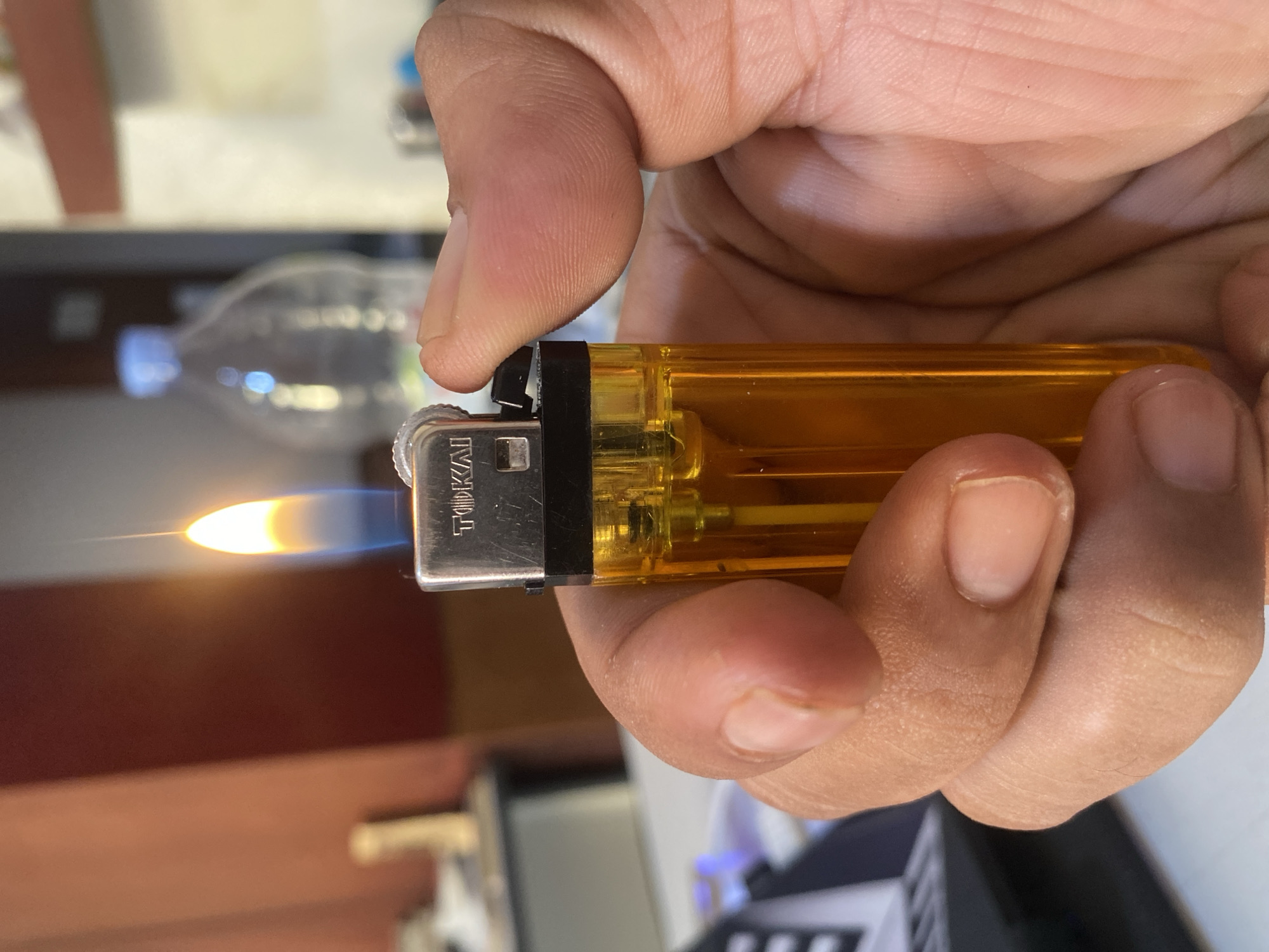 A close-up, high-angle shot shows a hand holding a clear, orange plastic lighter with a flame. The lighter has a silver metal top with the brand name "TIKAI" visible. The flame is bright yellow and blue, with a thin, tall, vertical peak. The hand is in the foreground, with fingers wrapped around the lighter, obscuring part of it. The background is out of focus, showing a blurry red wall on the left, and a plastic water bottle on a shelf in the upper right. The scene appears to be indoors, possibly in a residential or office setting. The image does not contain any information that could suggest a specific location in North Jakarta, Indonesia.