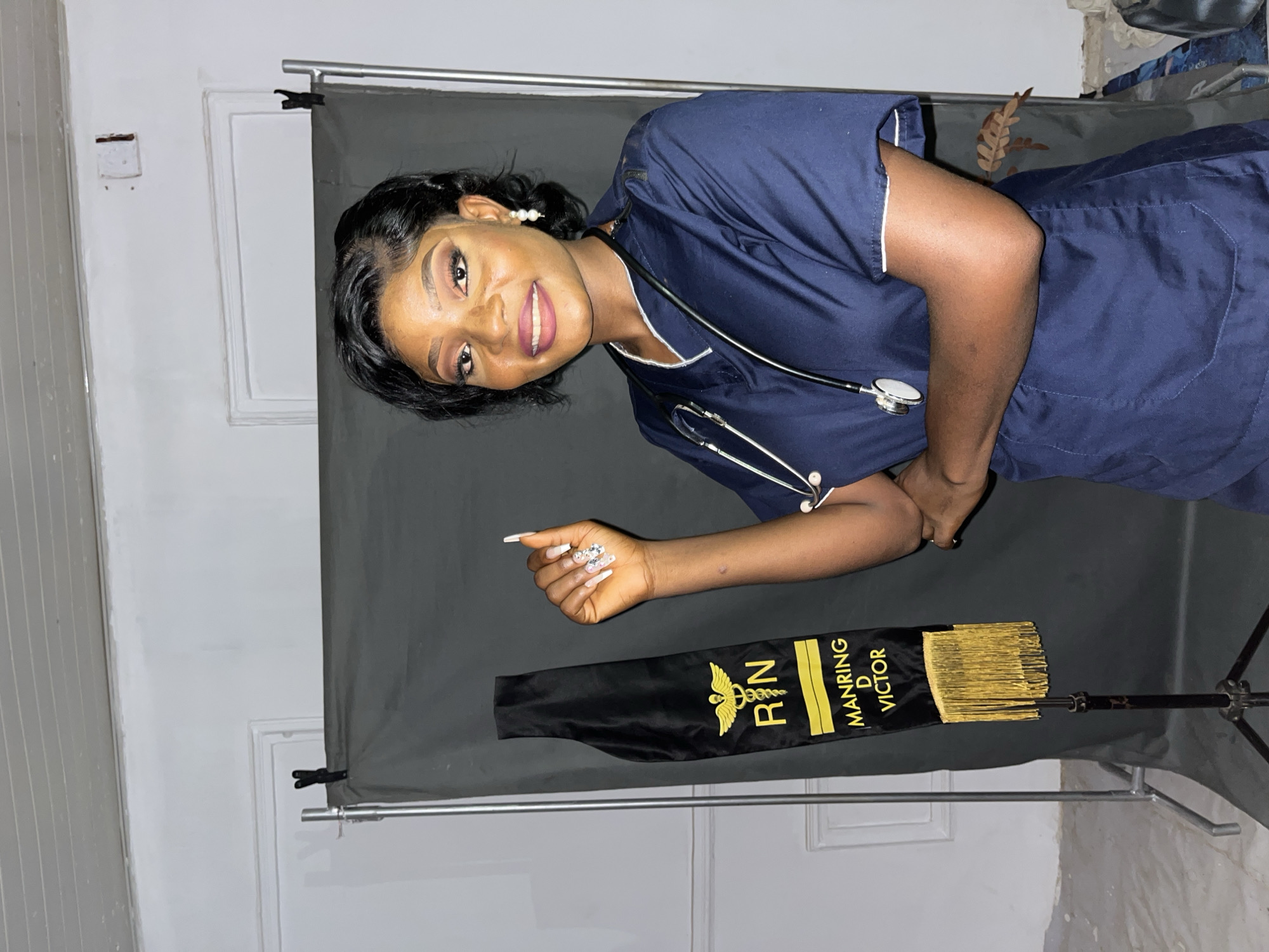 A woman in a navy-blue nursing uniform with a stethoscope around her neck is posing with a graduation sash. The black sash reads "RN" and "MANRING D VICTOR" in gold lettering and has gold tassels. The woman is smiling, and her right hand is extended. The background consists of a black backdrop and a dimly lit room with white walls. The location is Unknown, Unknown.