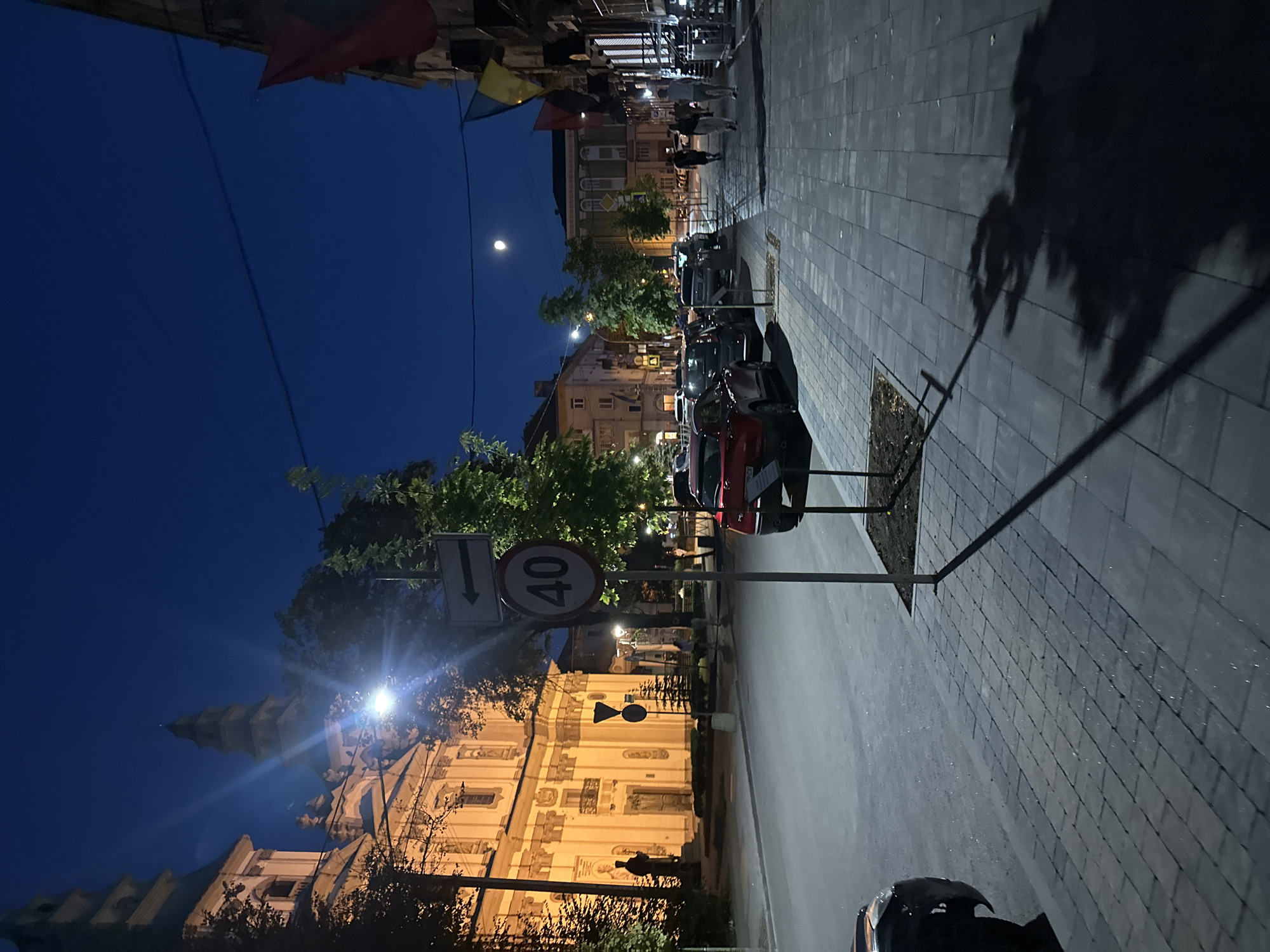 The image depicts a street scene at night in Ternopil, Ukraine, likely near a church or historical building. The sky is a deep blue, with a visible moon. The left side of the image features a grand, illuminated building with architectural details characteristic of a church or historical edifice, featuring a tall spire. A street lamp casts a bright light, creating a lens flare. Trees are present, casting shadows. A traffic sign indicating a speed limit of 40 is visible, along with a directional arrow sign. Several cars are parked along the street. The sidewalk is paved with stone tiles, and the street is asphalt. On the right side of the image, a building with flags (one appearing to be Ukrainian) is visible. A few individuals are walking on the sidewalk in the distance. The overall atmosphere is tranquil and illuminated by artificial lights.