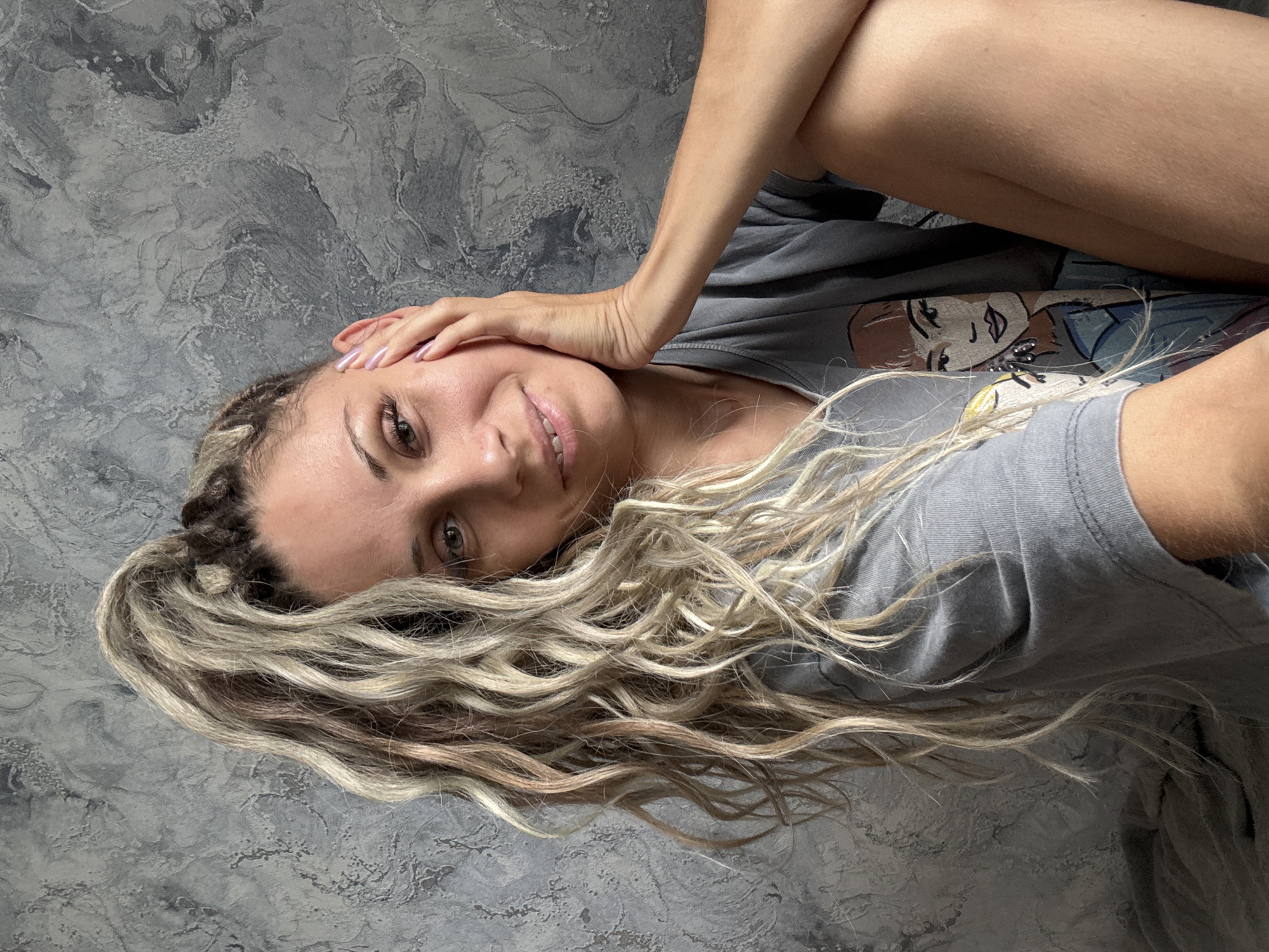 A portrait of a woman with blonde, curly dreadlocks pulled into a high ponytail against a textured grey wall. She is wearing a grey t-shirt with a graphic of a woman on it and is leaning her head on her hand. The image focuses on her upper body and face. The scene does not provide any information or visual cues to suggest its location in Rusanovo, Russia.