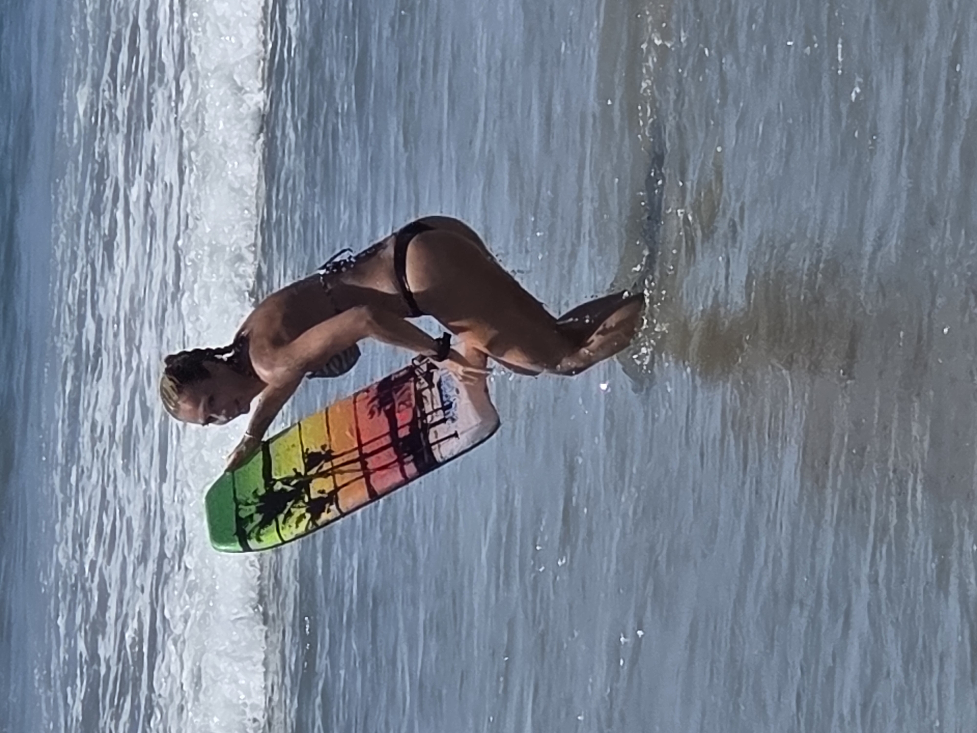 A woman in a bikini stands in the ocean water, holding a boogie board. The boogie board has a colorful design of palm trees against a sunset background. The woman is looking to her right, with her hair in a braid. The ocean has gentle waves, and the sky is bright and sunny. The scene appears to be on a beach in Santa Monica, California.
