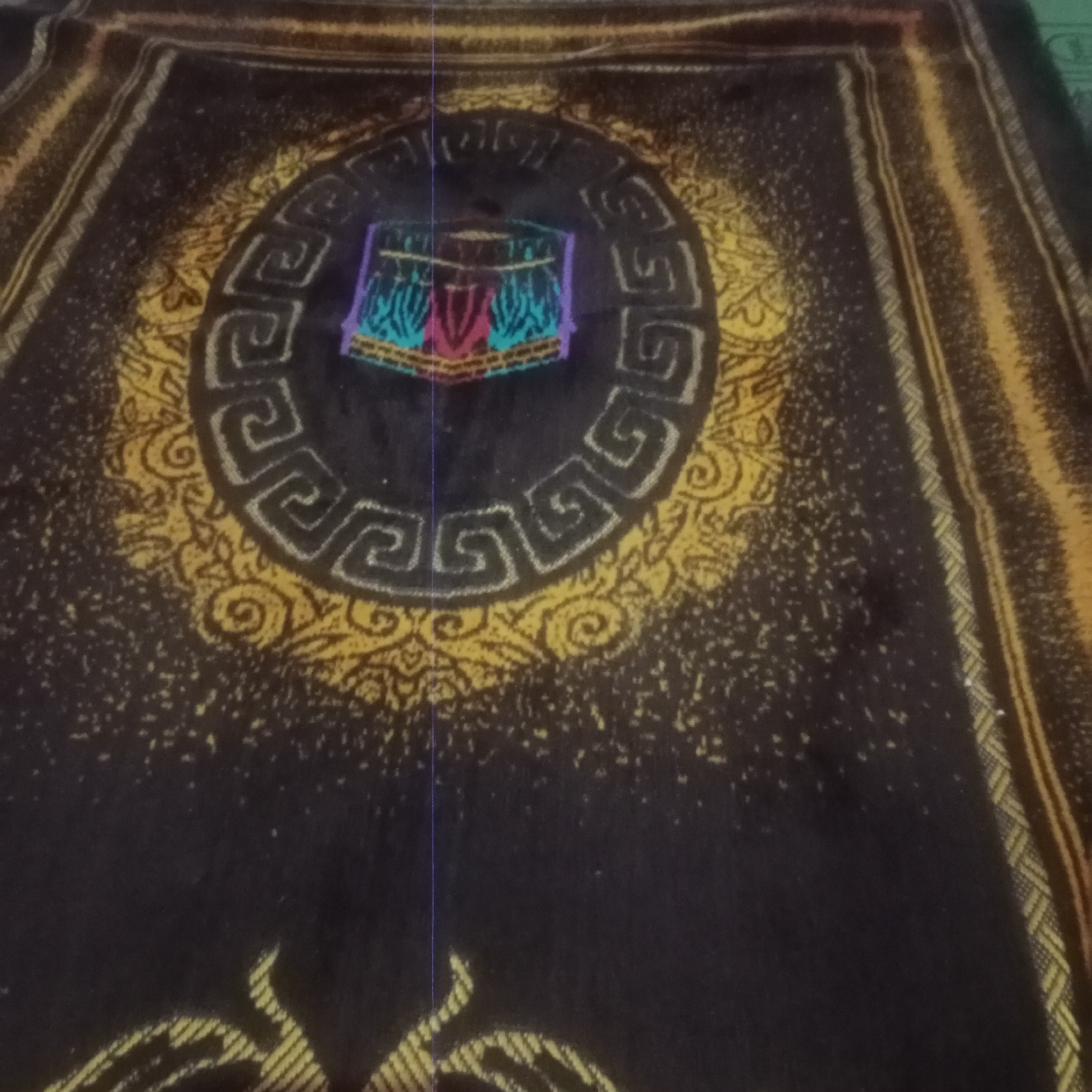 The image shows a close-up, overhead view of a prayer rug with intricate designs. The rug is predominantly black with gold and yellow patterns. In the center, there is a circular motif with Greek-key-like designs, framing a colorful depiction of the Kaaba in Mecca. The Kaaba is rendered in shades of purple, pink, turquoise, and green. The background of the rug is speckled with small, light-colored dots, possibly representing texture or a stylistic element. Along the sides of the rug, there are striped borders in shades of gold and yellow. The rug appears to be made of a soft material, likely fabric. The lighting in the image is somewhat dim, with a slight purplish cast, suggesting an indoor setting or low-light conditions. There is no indication of the city of Tegowanu Kulon, Indonesia, in the image itself. The image focuses solely on the prayer rug.