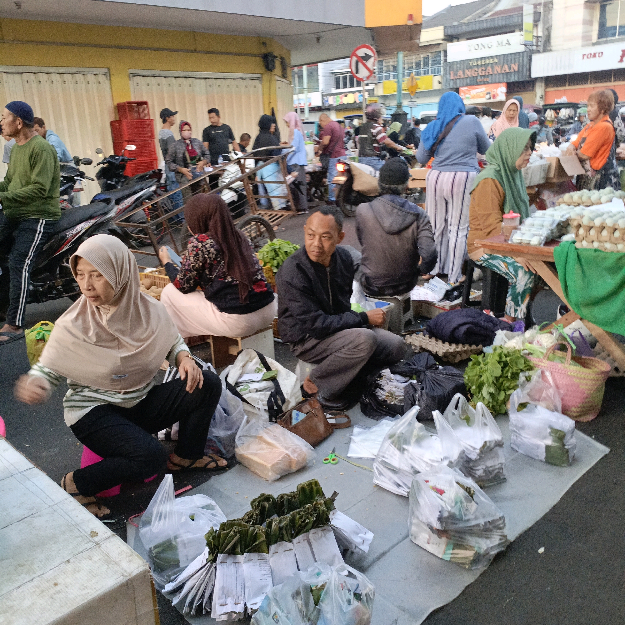 The image depicts a street scene in Salatiga, Indonesia, likely a market area. In the foreground, a woman wearing a beige hijab and a striped shirt is crouched down, looking to her left with a focused expression. She is dressed in black pants and sandals. Beside her, to the left, is a large light-colored box and several plastic bags containing goods, including what appear to be packaged items. To the right of the woman, there are more plastic bags and a pink woven basket.
Further back in the scene, a man in a black jacket and grey pants is seated on a small stool, looking to his right. He appears to be a vendor. Behind him, there are more packaged items and various goods on display, including what look like rows of eggs in cartons. A green cloth is draped over a table or display.
The background shows a bustling street with people, motorcycles, and buildings. Some individuals are wearing hijabs and traditional Indonesian clothing. There are signs in the background, including one with Indonesian text that reads "YONG MA" and "TOKO". A traffic sign with a red circle and a diagonal line is visible above the street. The overall impression is of a lively street market environment.