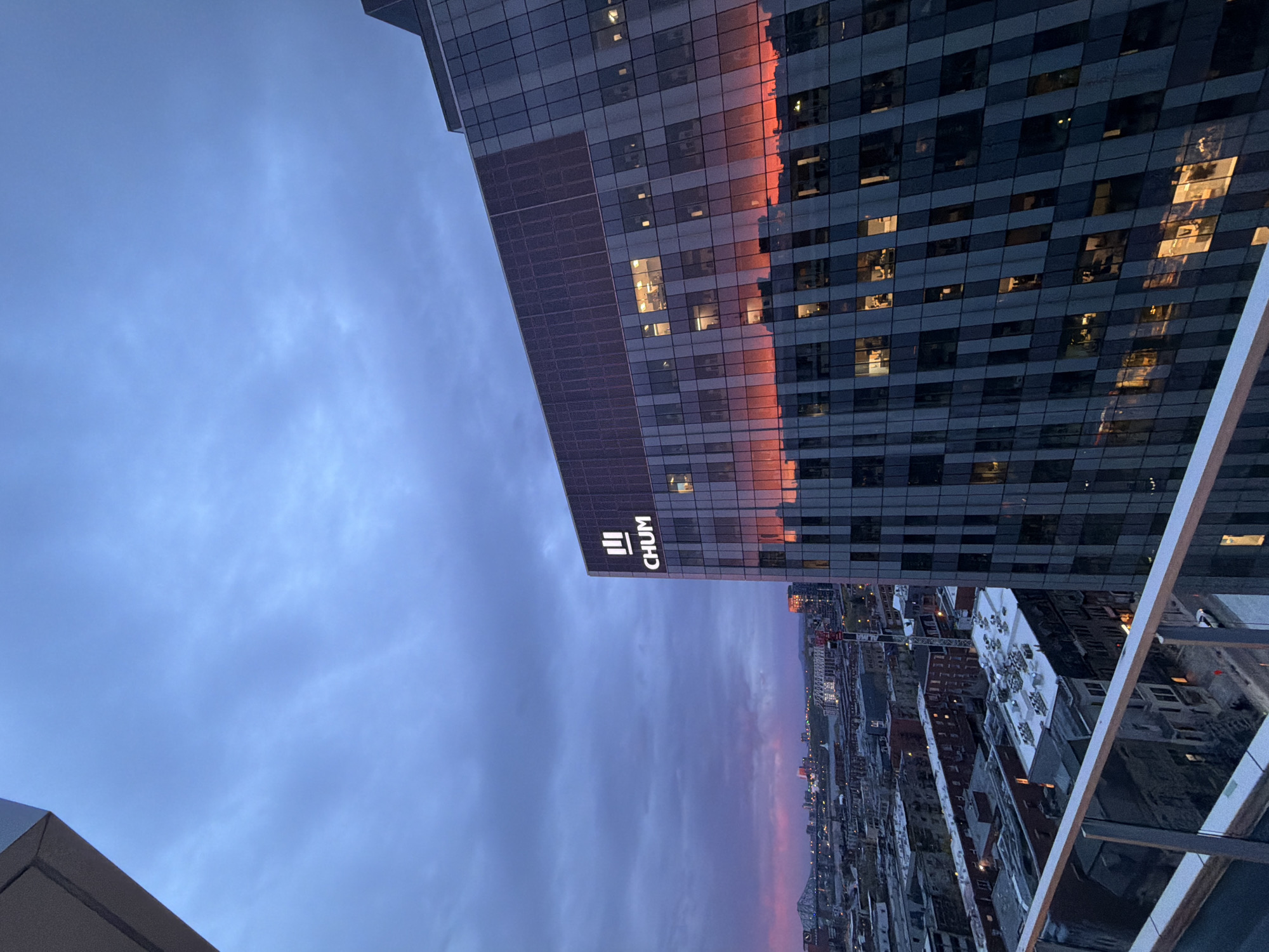 The image presents an upward view in Montreal, Canada, likely taken from a high vantage point such as a rooftop or high-rise balcony. The sky, taking up a large portion of the frame, displays a mix of blue and pink hues, indicating either sunrise or sunset. Prominently featured is the CHUM (Centre hospitalier de l'Université de Montréal) building. The lower part of the image includes a cityscape with various buildings and urban structures, with a pinkish color washing over the city from the sunset/sunrise. Elements of the rooftop or balcony from where the image was taken are visible in the bottom corners.