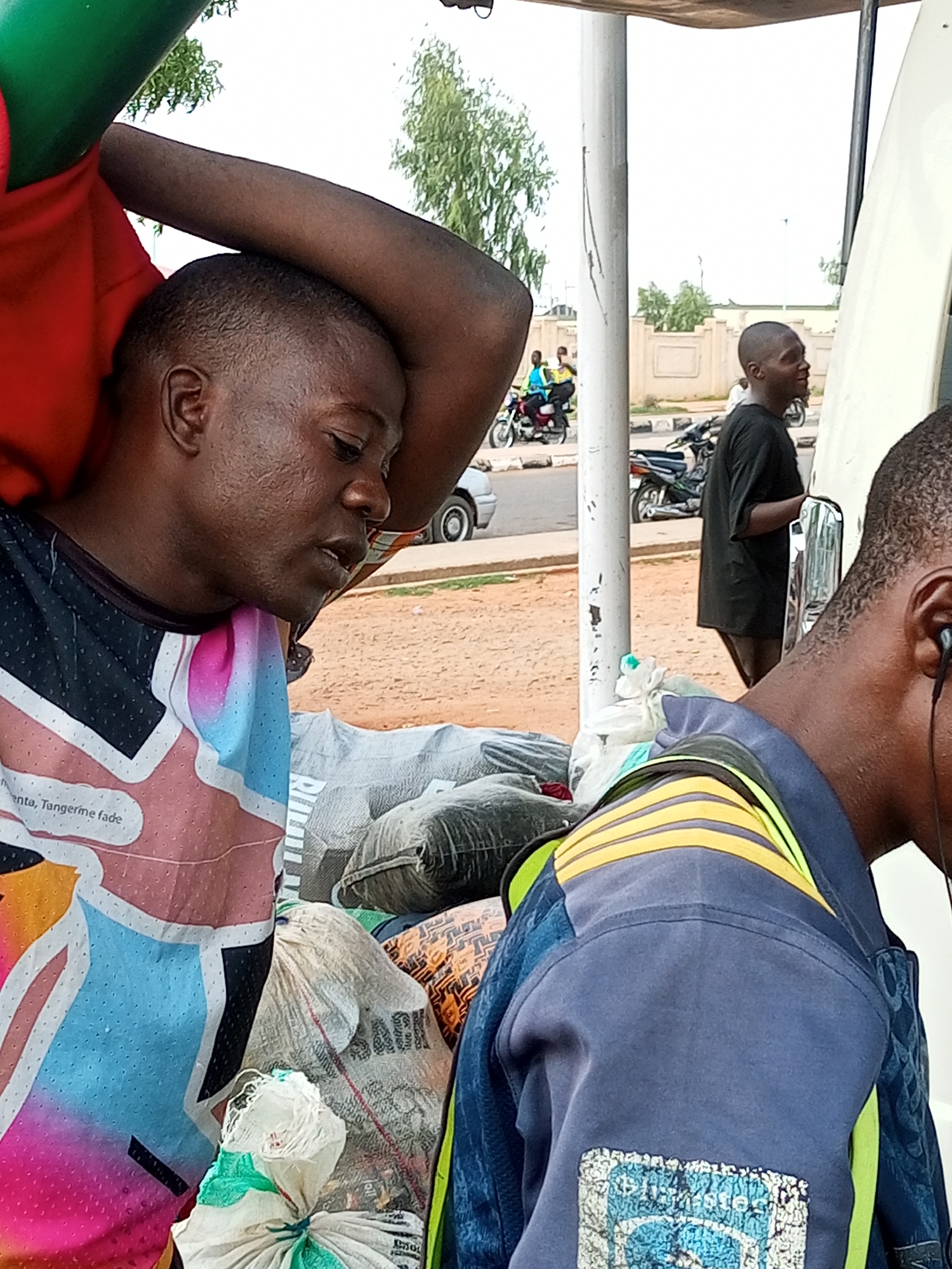 The image captures a scene on a street in Gombe, Nigeria, likely a commercial area due to the presence of vehicles and people. In the foreground, a Black man is seated, with his right arm resting on his head and his left arm bent upwards. He is wearing a brightly colored shirt with a graphic design that includes pink, blue, and yellow elements. His expression appears thoughtful or tired.
Behind him, another Black man is standing, wearing a black t-shirt. He is looking to his right, away from the camera. A white van or bus is partially visible on the right side of the frame. In the background, several motorcycles are parked and in motion, suggesting a busy thoroughfare. One motorcycle carries two individuals. There are also several bags, some appearing to be sacks, piled up near the seated man, possibly indicating goods for sale or transport. The environment appears to be outdoors, with a dusty or sandy ground and a bright sky, suggesting a warm climate. The overall impression is one of everyday life and activity in a Nigerian city.