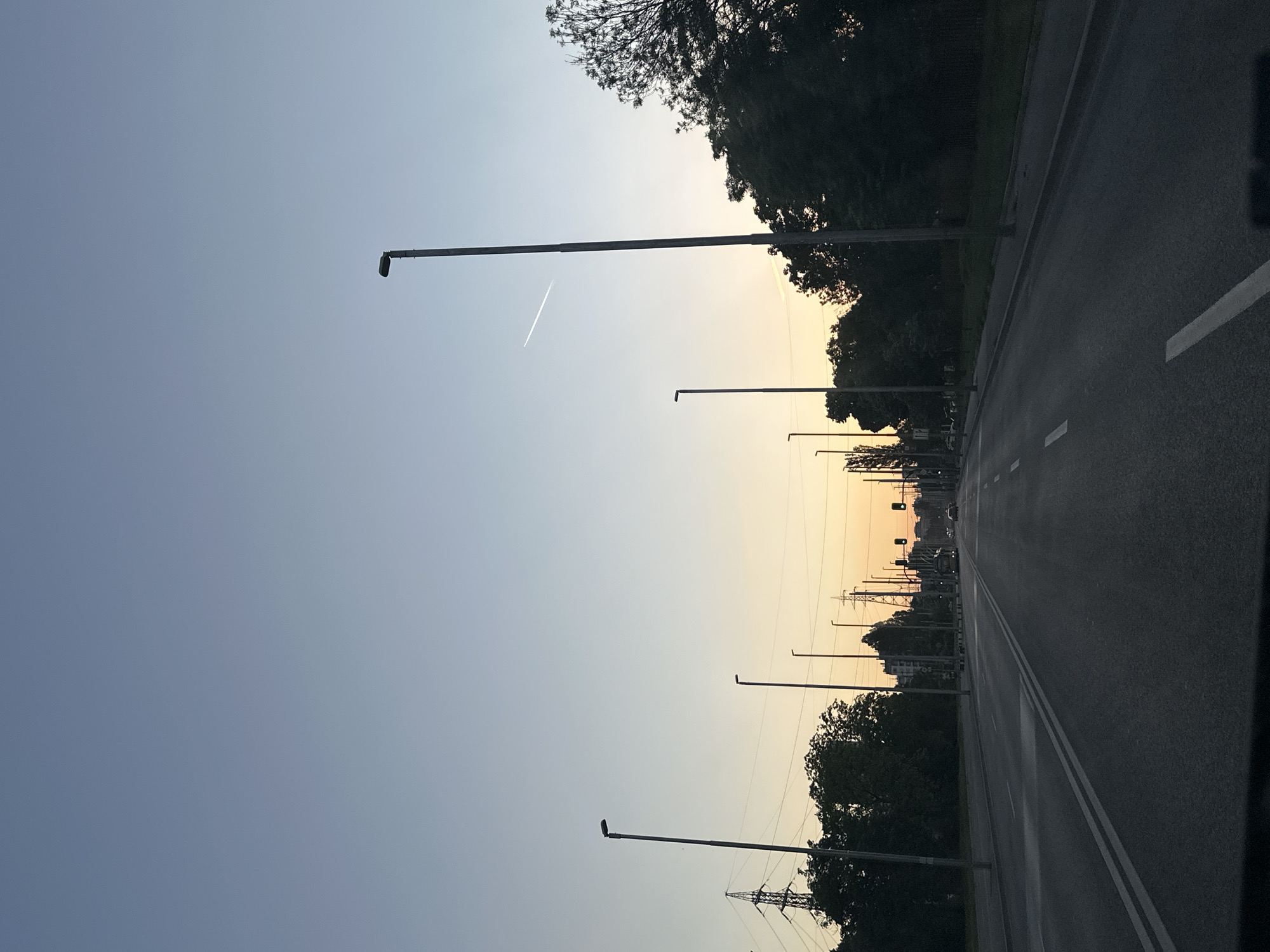 A slightly angled photograph shows a straight asphalt road receding into the distance. Trees flank the right side of the road, with rows of street lamps lining both sides. Overhead power lines are visible among the trees. The sky transitions from a clear blue to a light orange near the horizon. A faint contrail from an airplane is visible high in the sky. The scene appears to be near the Perlovska River near the bridge "Mosta na Vлюбeните", Sofia, Bulgaria.