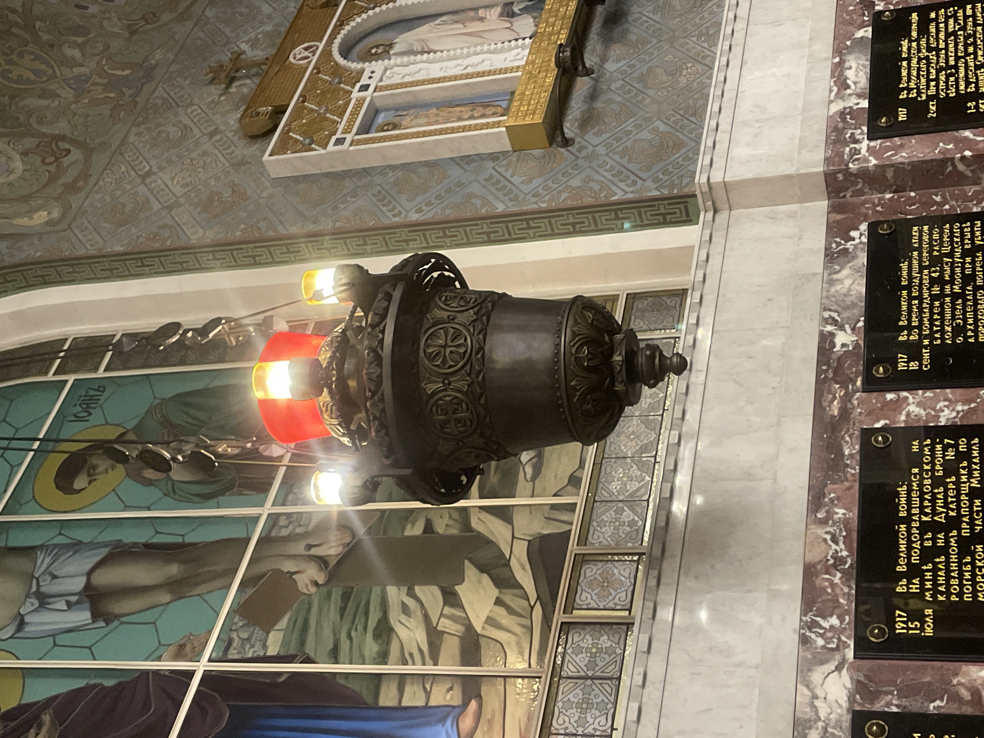 The image depicts the interior of a building, likely a church or memorial, featuring stained glass windows and ornate decor. A large, dark-colored hanging lamp with red and yellow lights is prominently displayed in the foreground, casting a warm glow. Behind the lamp, a stained glass window depicts a religious scene, possibly the crucifixion of Jesus, with figures and symbolic imagery. To the right, a religious icon or painting is mounted on a wall adorned with patterned wallpaper. Below the stained glass, a marble-like surface forms a base, upon which black plaques with inscriptions are mounted. These plaques appear to be memorials, with dates and text written in Cyrillic script. The overall atmosphere suggests a place of historical significance and religious devotion. This scene is located in Kronstadt, Russia, specifically within the Naval Cathedral of Saint Nicholas.