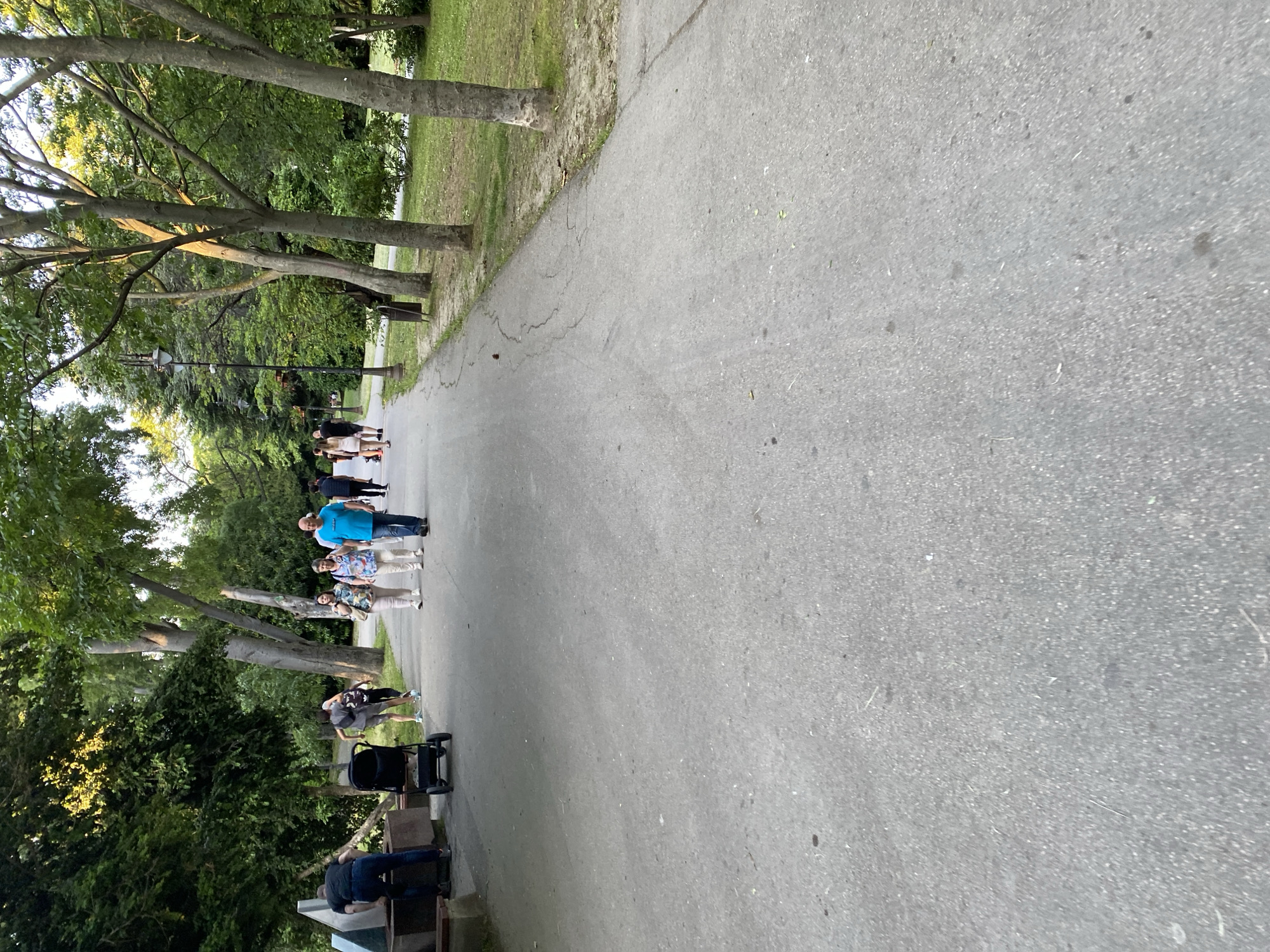 The scene is a paved pathway through a park or green space, lined with trees on both sides. A group of people are walking along the pathway, including one person wearing a blue shirt. There is a stroller and some benches visible on the side of the path. The location is the city of Varna, Bulgaria.