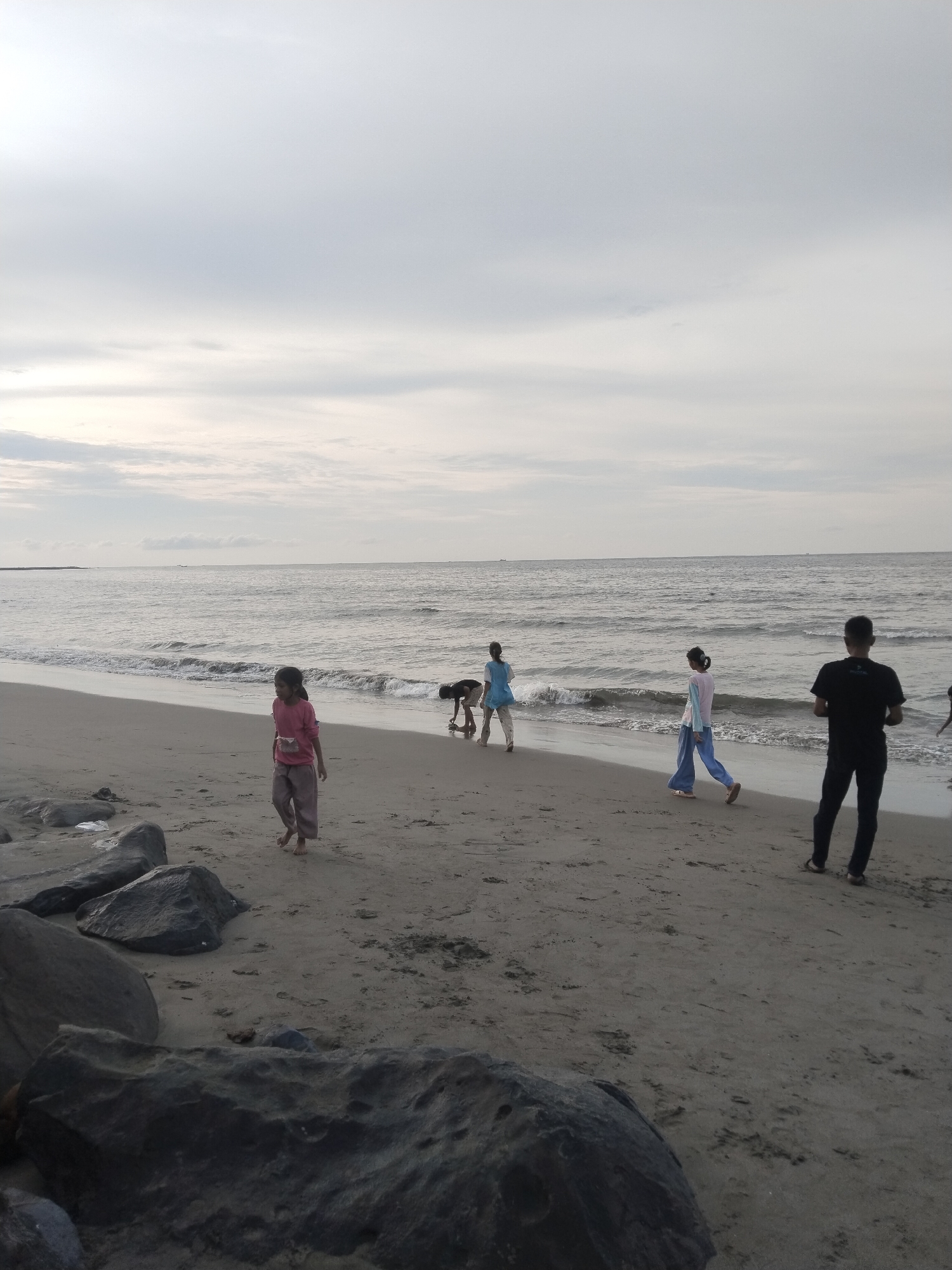The image depicts a beach scene in Lhokseumawe, Indonesia. The sky is overcast with clouds, and the sea is calm with gentle waves. The sand is a light brown color, and there are large rocks scattered along the shore, particularly in the foreground.
Several people are present on the beach. In the middle ground, a young girl wearing a pink top and light-colored pants walks towards the left of the frame. To her right, a child wearing a blue shirt is bent over, possibly playing or looking for something on the sand. Further to the right, two figures, one in a pastel-colored top and blue pants, and another in a black t-shirt and dark pants, are walking along the water's edge. The individual in the black t-shirt is facing away from the camera.
The environment suggests a casual day at the beach, with the presence of both children and adults. The overall atmosphere is subdued due to the cloudy weather. The rocks in the foreground and the expanse of sand and sea are characteristic of a coastal environment.